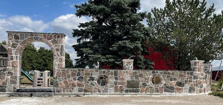 A long stone wall with an arched entrance stands in front of a red barn with pine trees in front of it