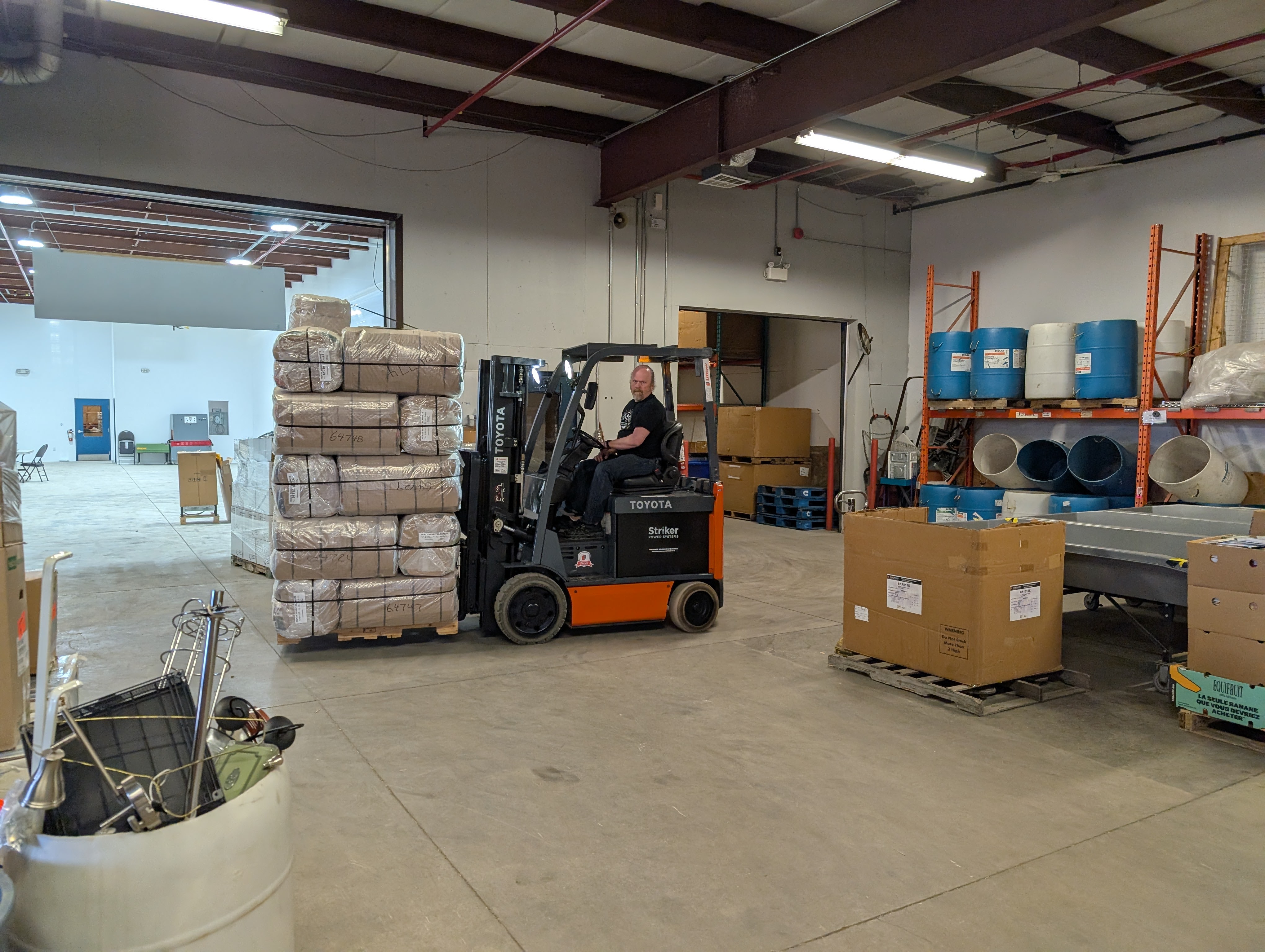 A man driving a fork lift carrying kits in a warehouse.