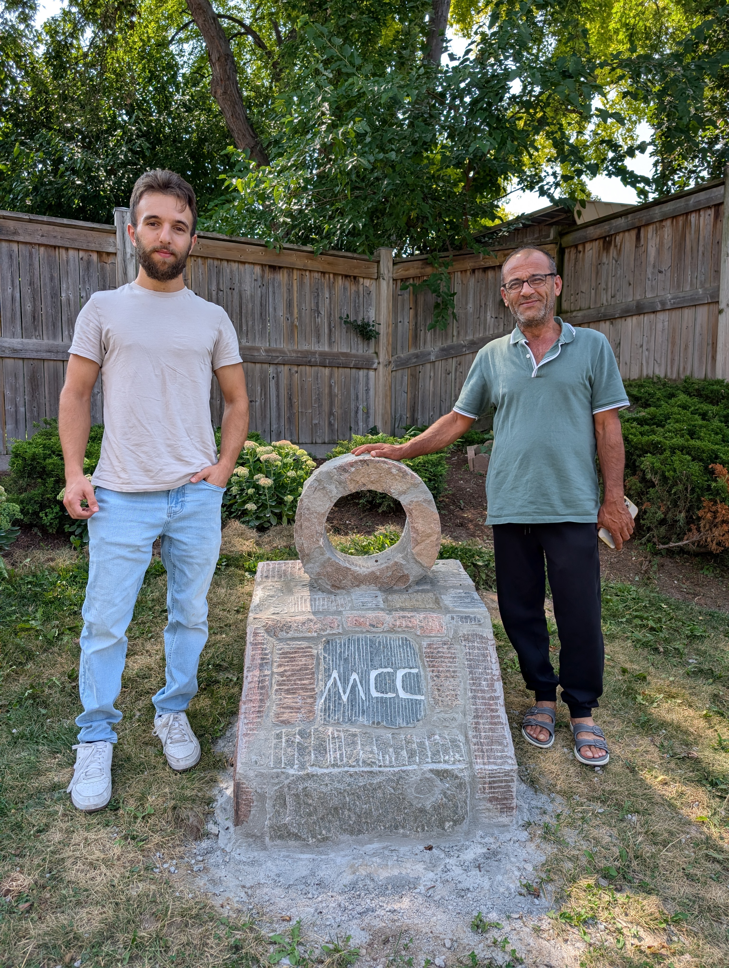 A young man and a middle-aged man stand next to a stone monument with the words MCC carved into it. The older man’s hand is resting on a stone circle on top of the monument.