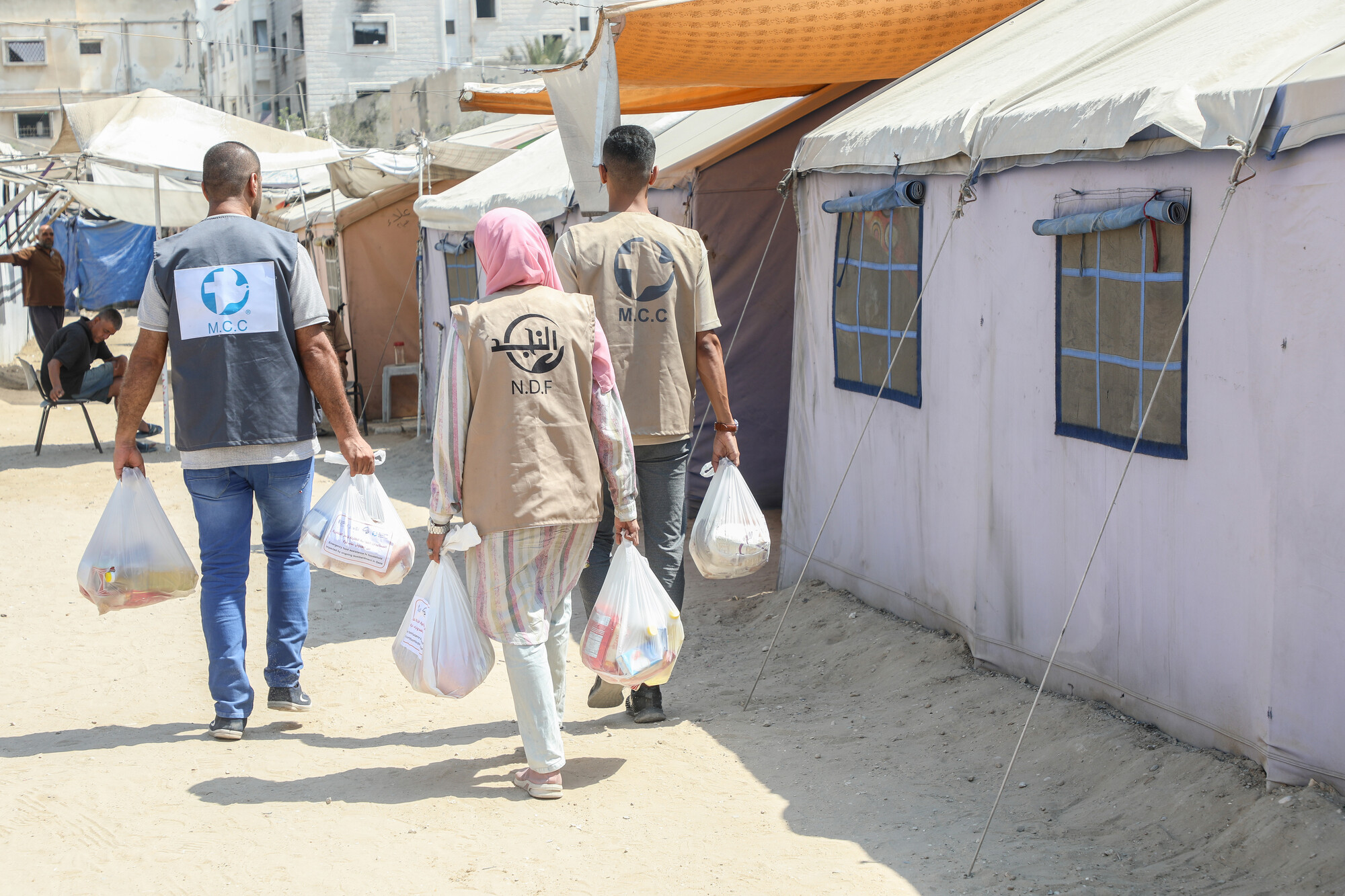 Three individuals walk towards tents, carrying bags. Two wear vests with the MCC logo, while another has a headscarf. Tents are in the background.