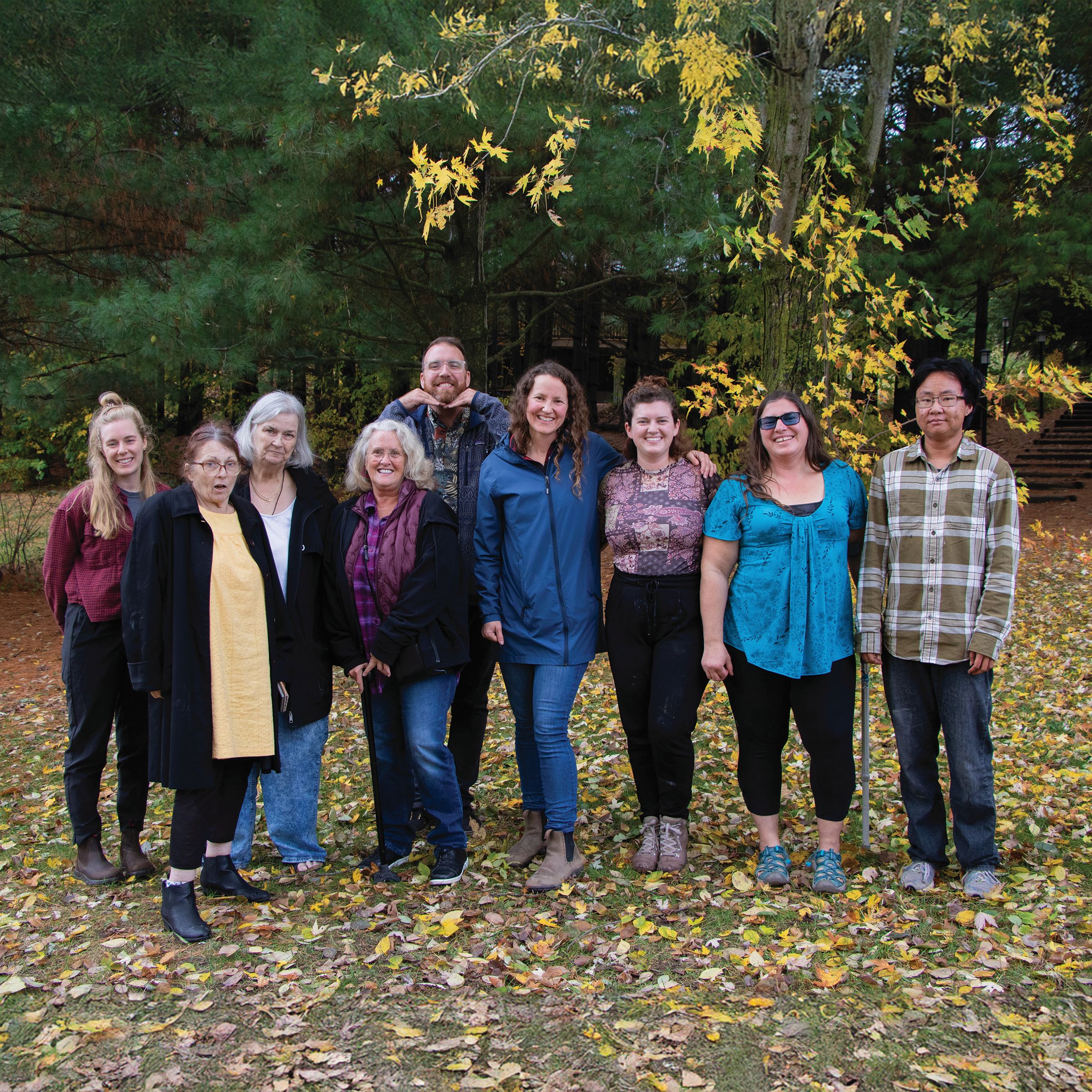 Nine people stand smiling, facing the camera in an autumnal forest.