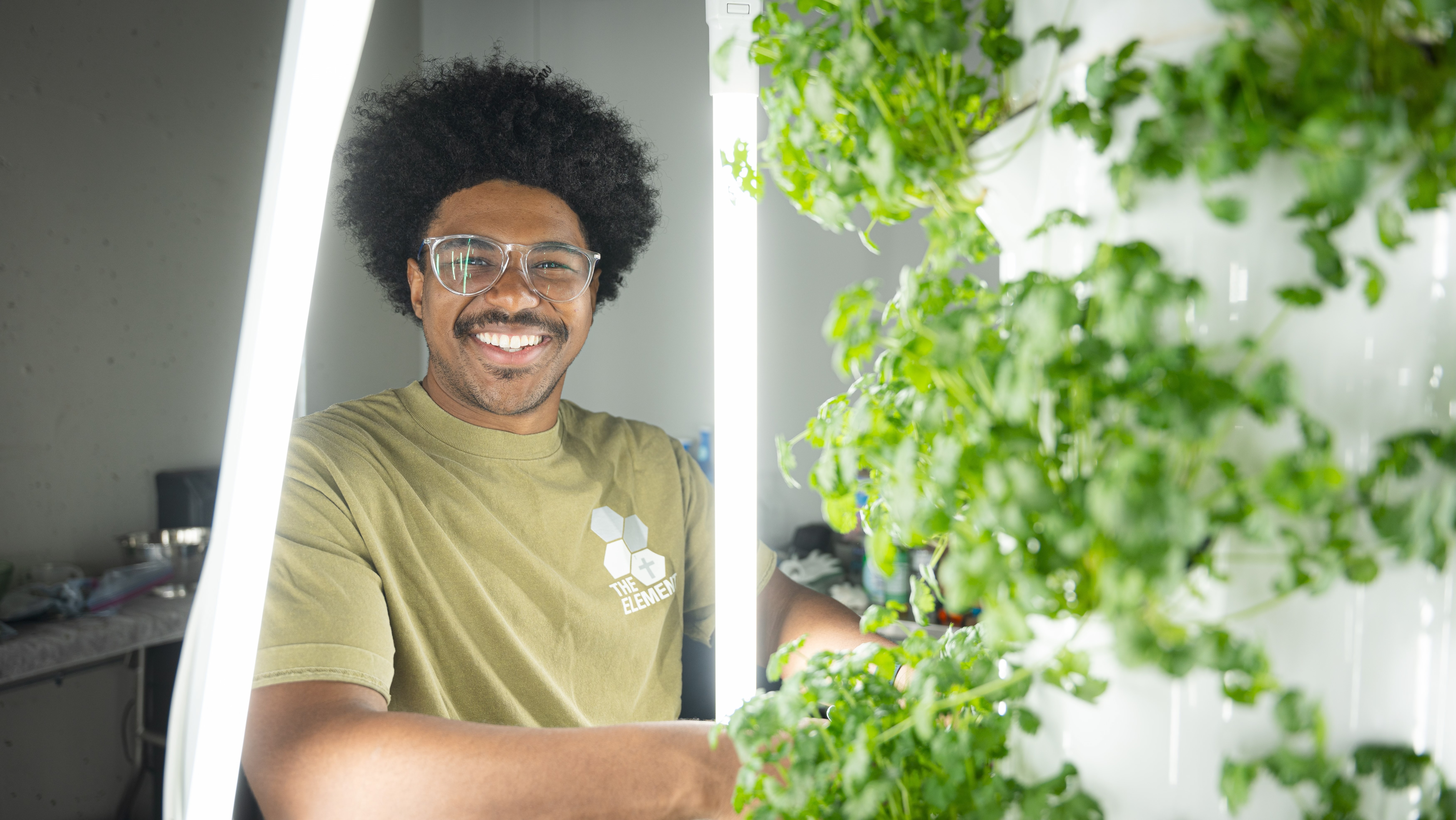 man smiles at camera as he tends to green plants growing in a vertical tower