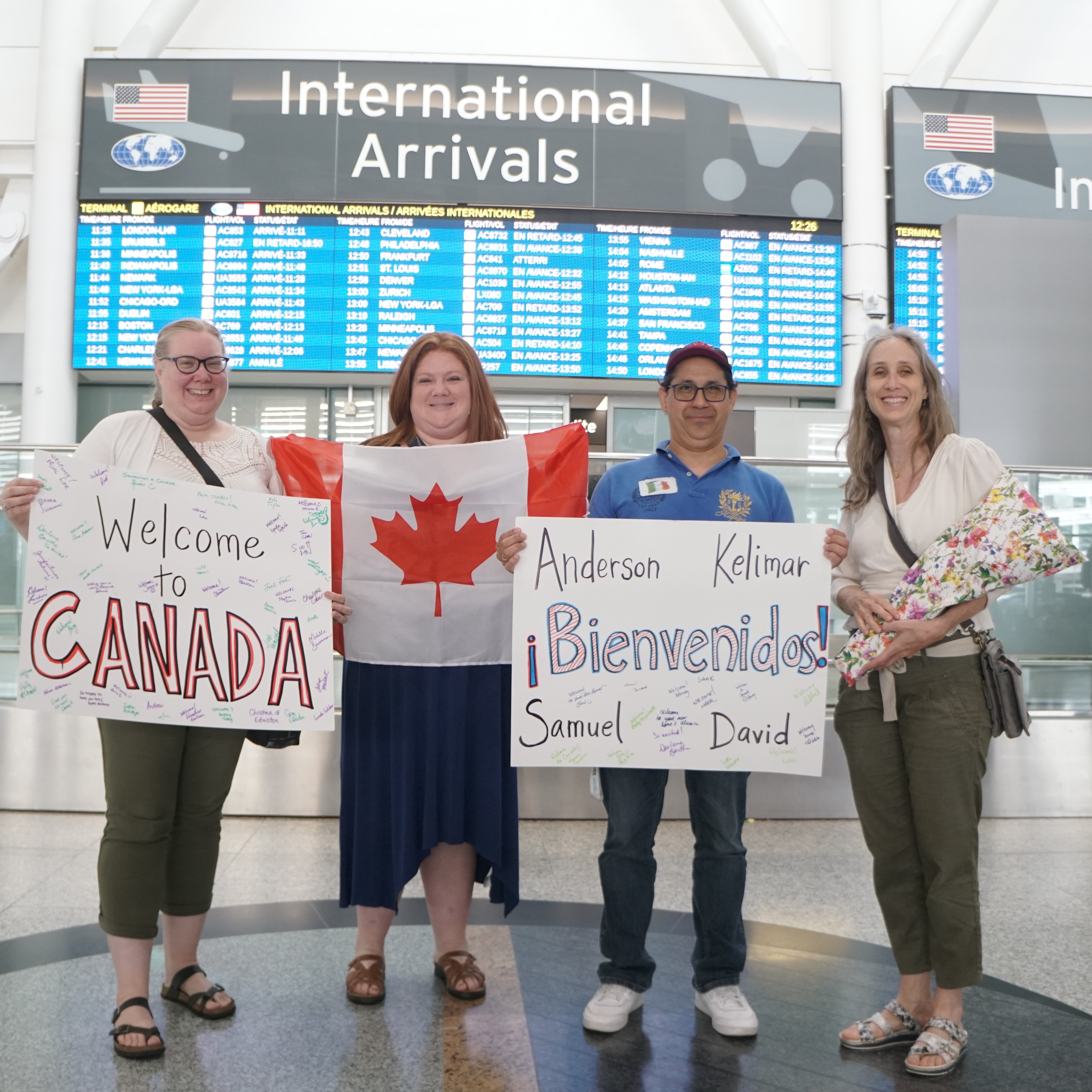 Four people stand in front of an arrivals board at an airport, holding handmade signs and gifts that express welcome to Canada.