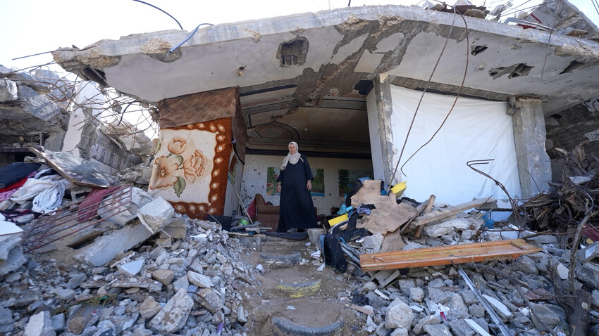 A woman stands amidst rubble and debris, with a partially collapsed building and scattered belongings around her.