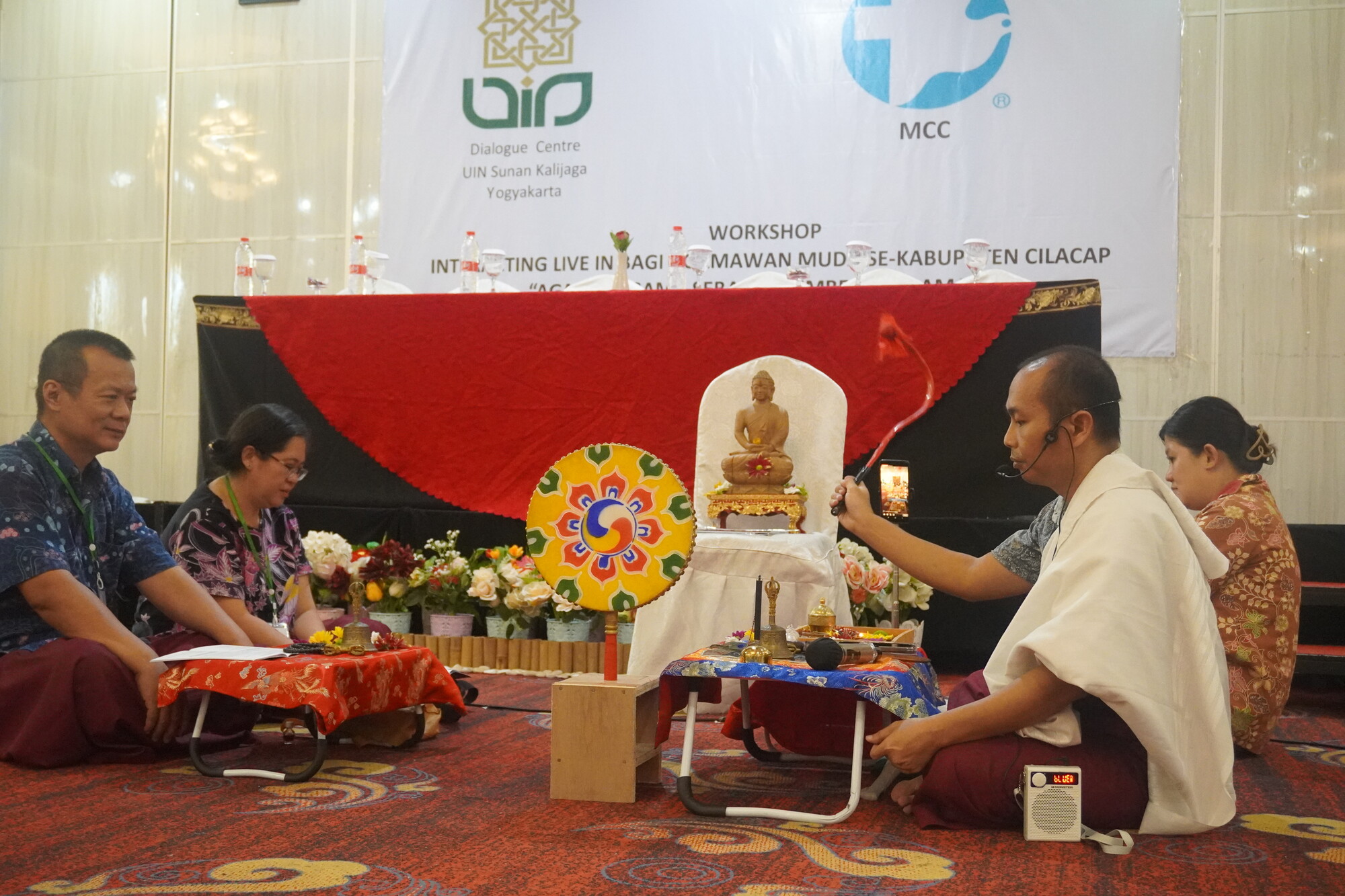 A workshop setting with participants seated around a small altar featuring a statue, surrounded by floral arrangements.