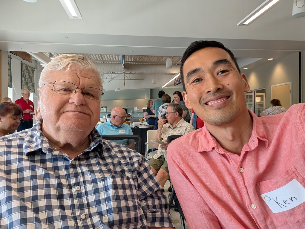 Two people sit next to each other and smile at the camera. One wears a nametag that says "Ken."