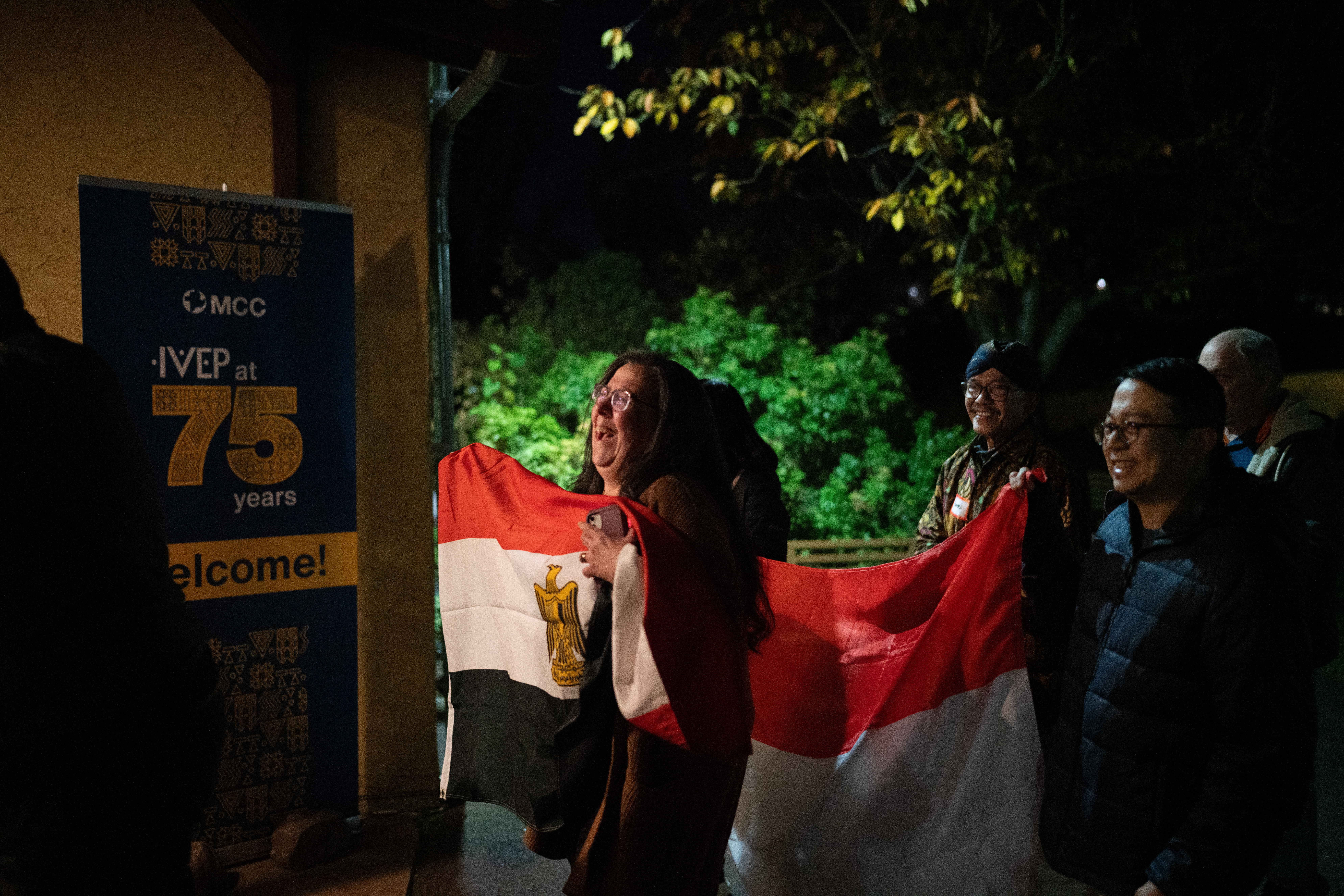 A smiling group carrying various international flags walk into a building with a welcome sign that reads, "IVEP at 75 years."
