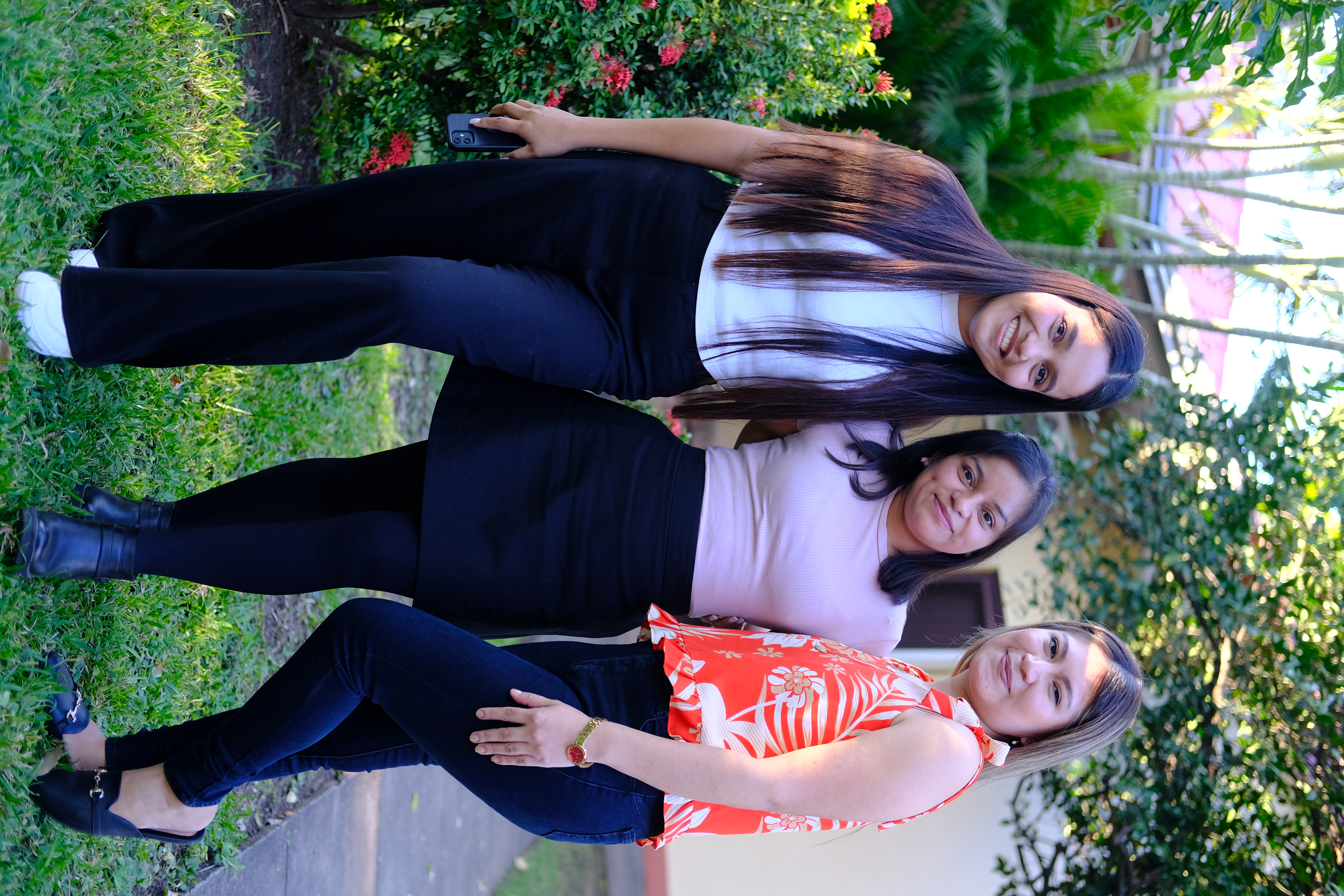 Three women stand together outdoors, smiling. They are dressed in various styles, with greenery and flowers in the background.