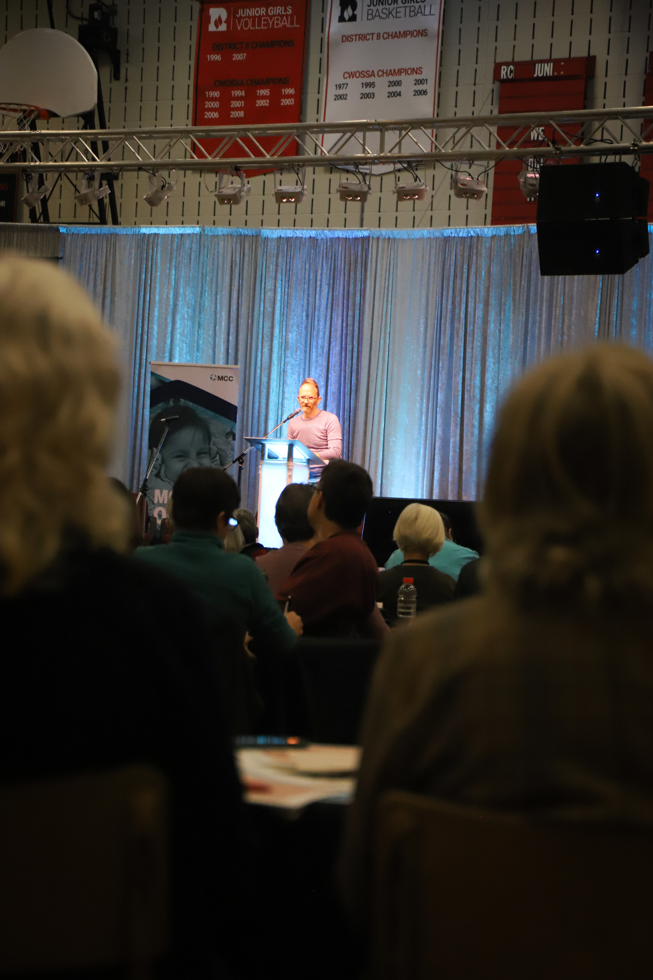 A man speaks at a podium with spectators in shadow in the foreground.
