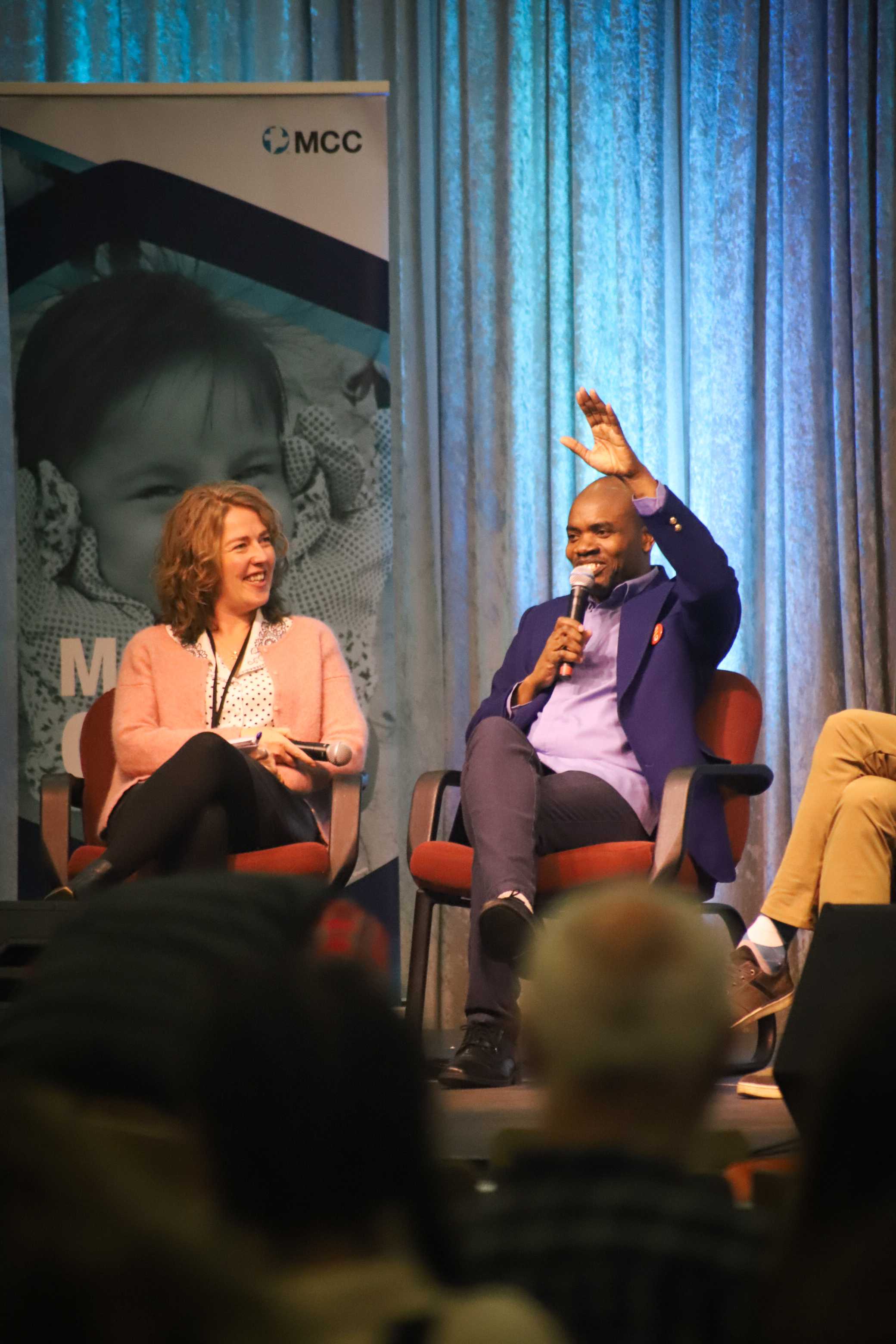 A woman and man sit on a stage with people watching; the man is smiling and holding his hand in the air.