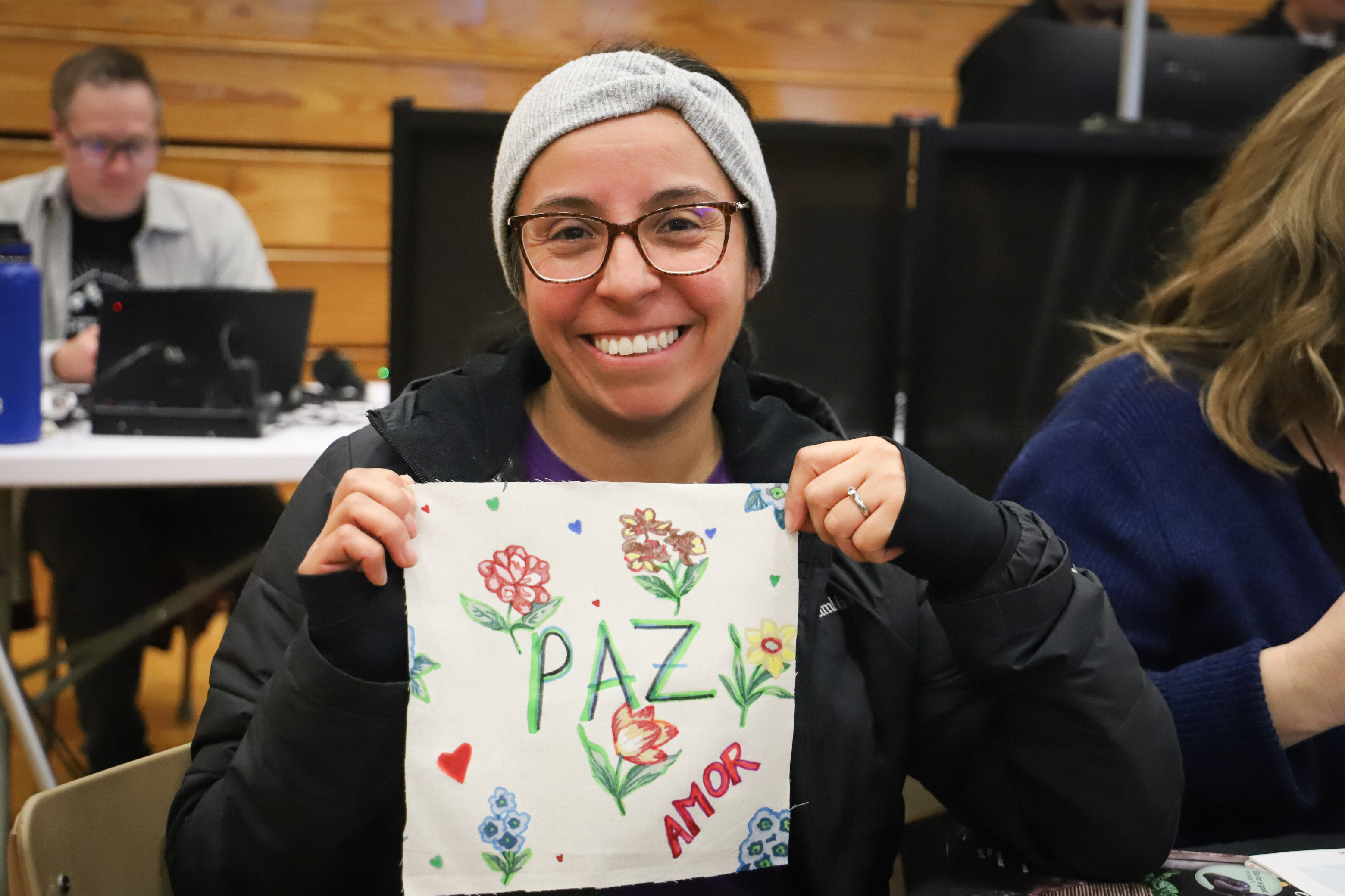 A smiling woman with glasses and dark hair holds up a square piece of cloth with the words "Paz" and "amor" on it