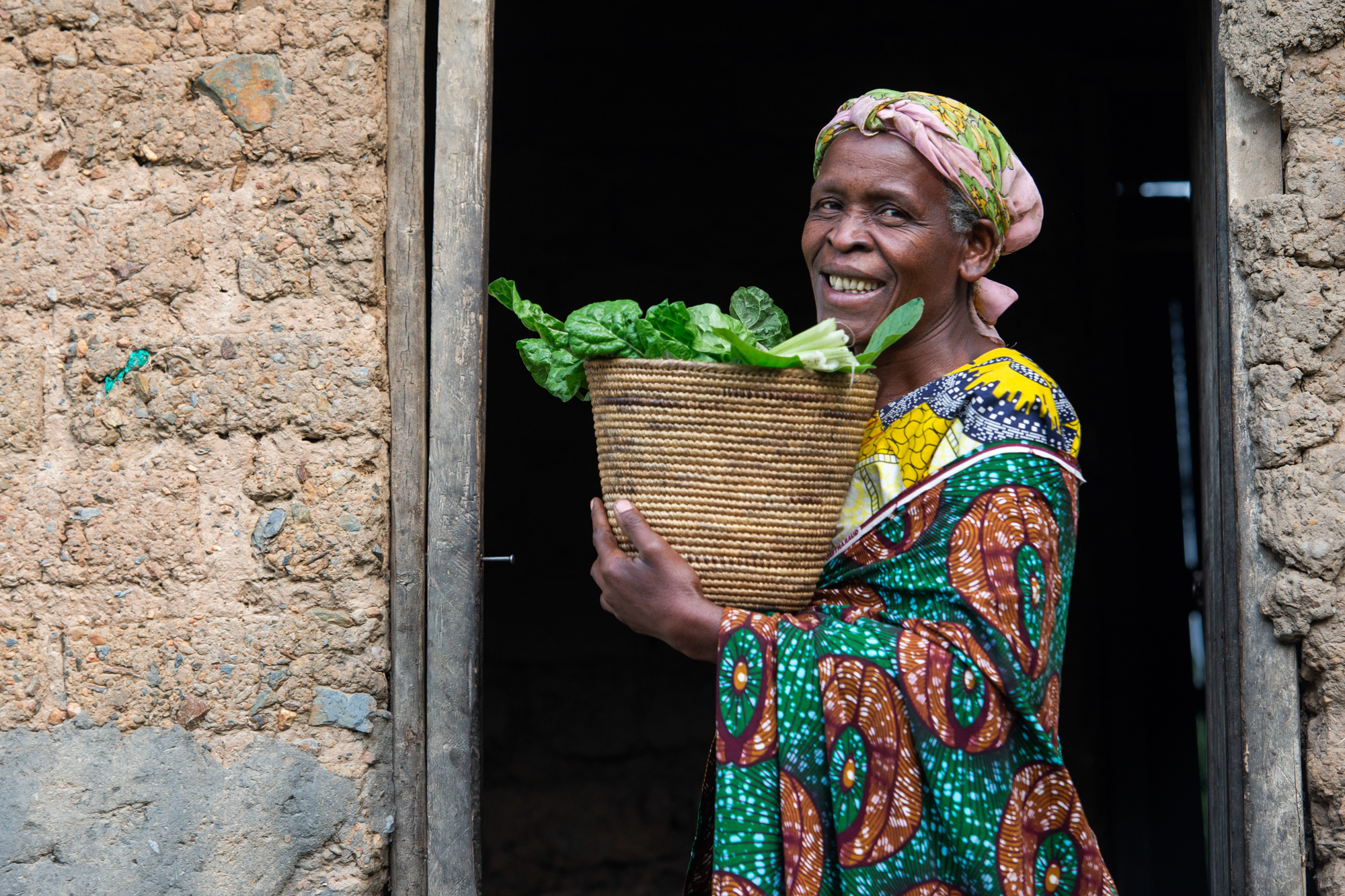 A smiling woman in colorful attire stands in a doorway holding a basket of leafy greens. The building appears to be made of mud bricks.