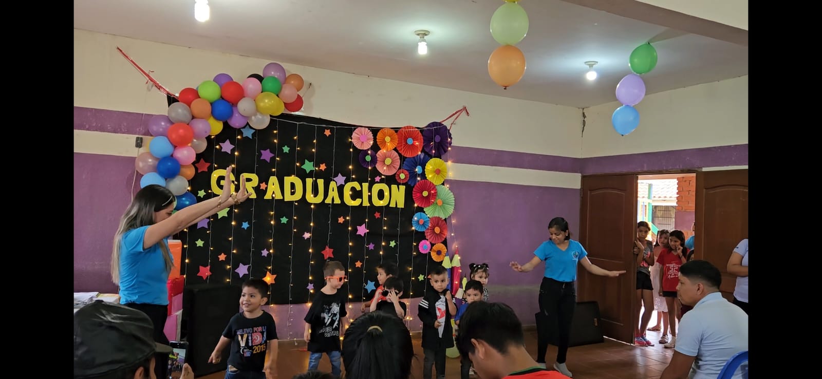 A decorated room with balloons, a "Graduación" banner, children performing, and adults watching from seated positions.