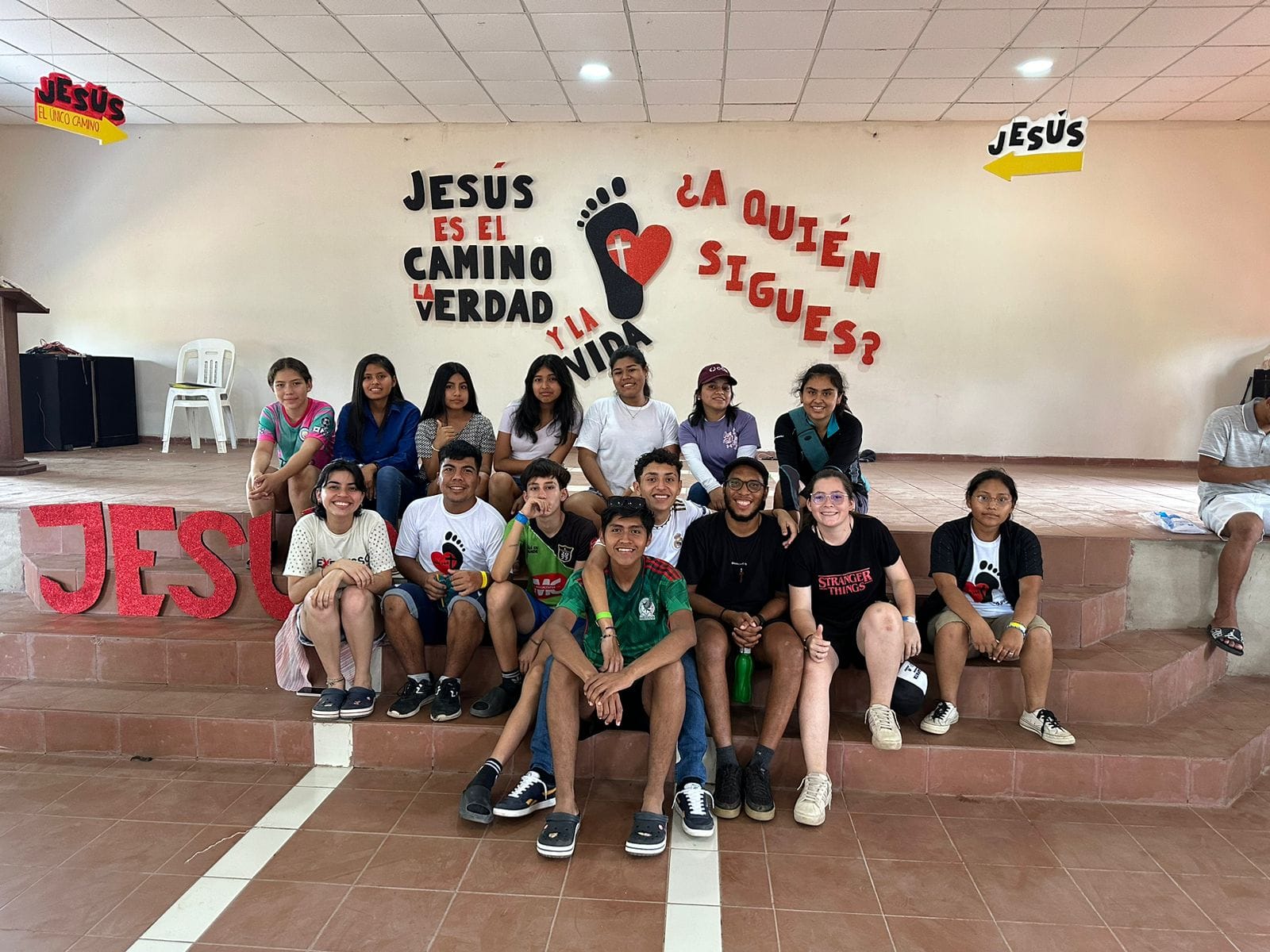 A group of young people sits on steps in a room with a stage. There are banners and signs with text related to Jesus.