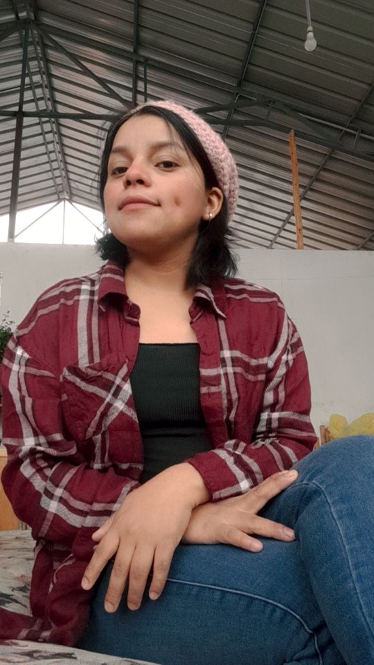 A person sits with a relaxed posture, wearing a plaid shirt and a beanie, against a backdrop of a simple indoor setting.
