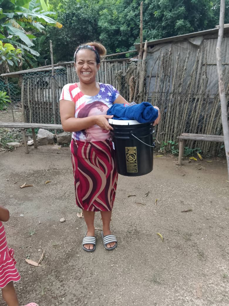 A smiling woman stands outdoors holding a bucket with clothes. She wears a striped skirt and a star-patterned top. A child is partially visible on the left, and there's a wooden fence behind.