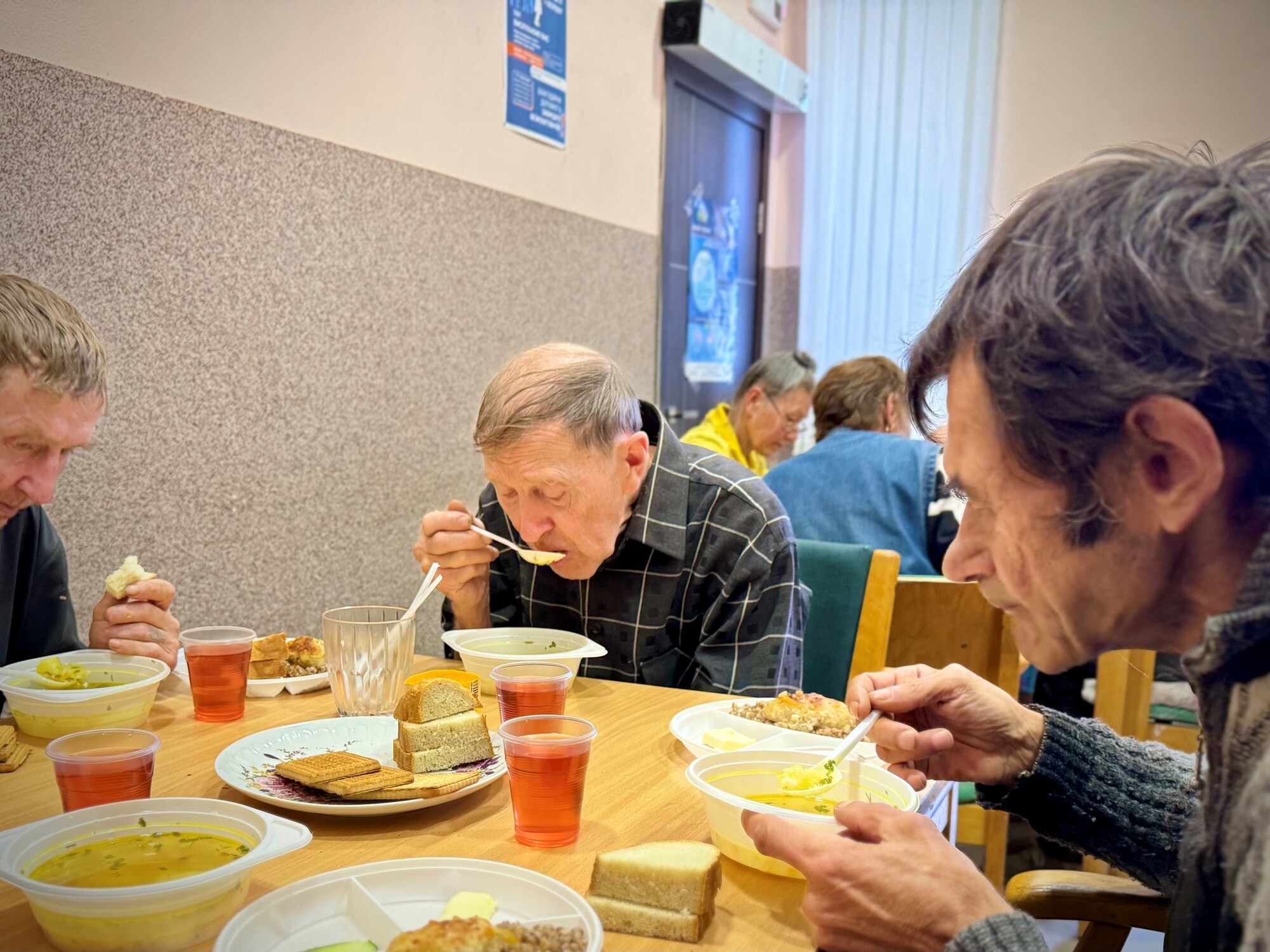 + The photograph shows several people seated at a table, eating soup and bread, with plates and glasses in front of them.