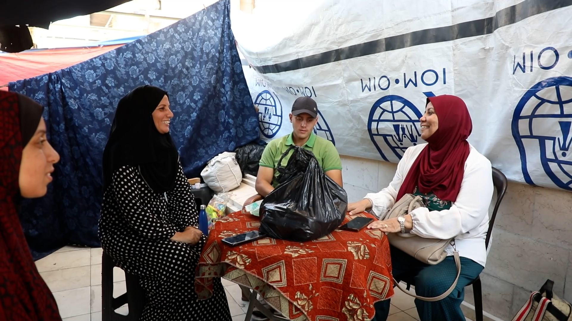 A women with a red head covering sits at a table with a man in a green shirt and a women in a white and black dress with a black head covering.