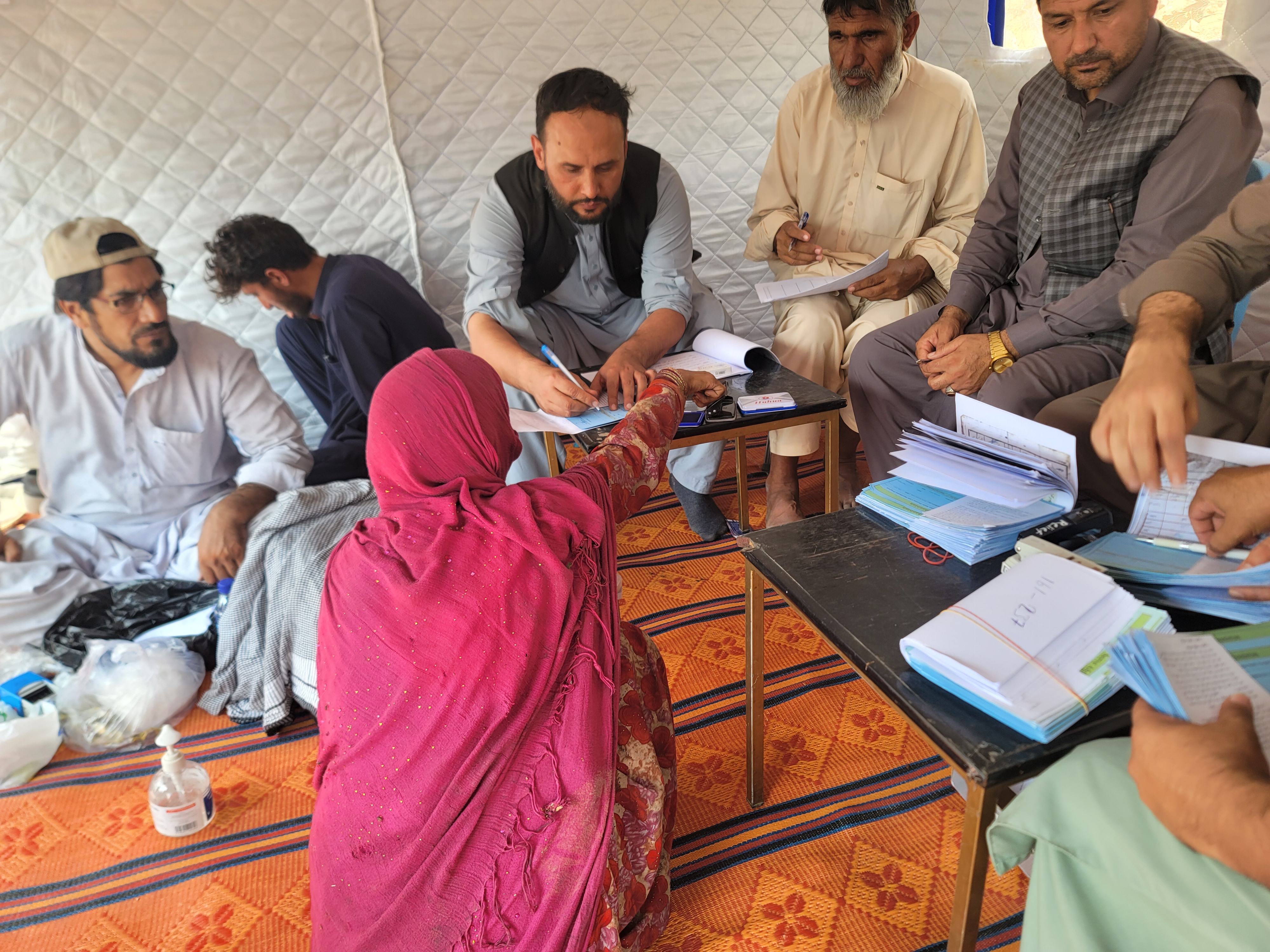 A group of people sits around a table in a tent, engaged in discussion and paperwork, with one person kneeling in front.