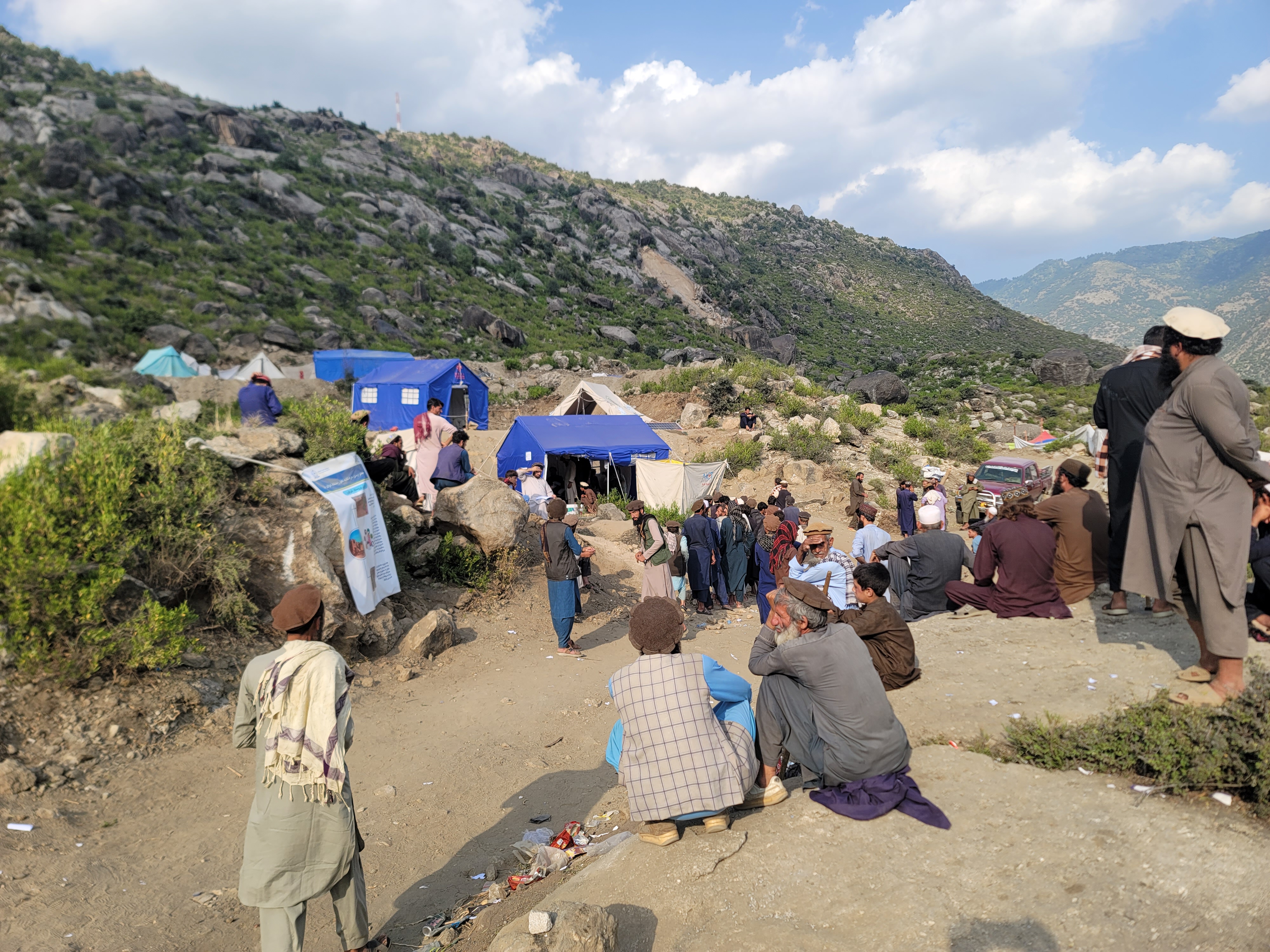  The photograph shows a mountainous landscape with tents, people gathered, and rocky terrain. Some individuals are seated or standing.