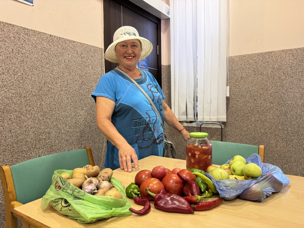A woman stands beside a table filled with various fruits and vegetables, including potatoes, apples, and peppers, in a room with a door and windows.