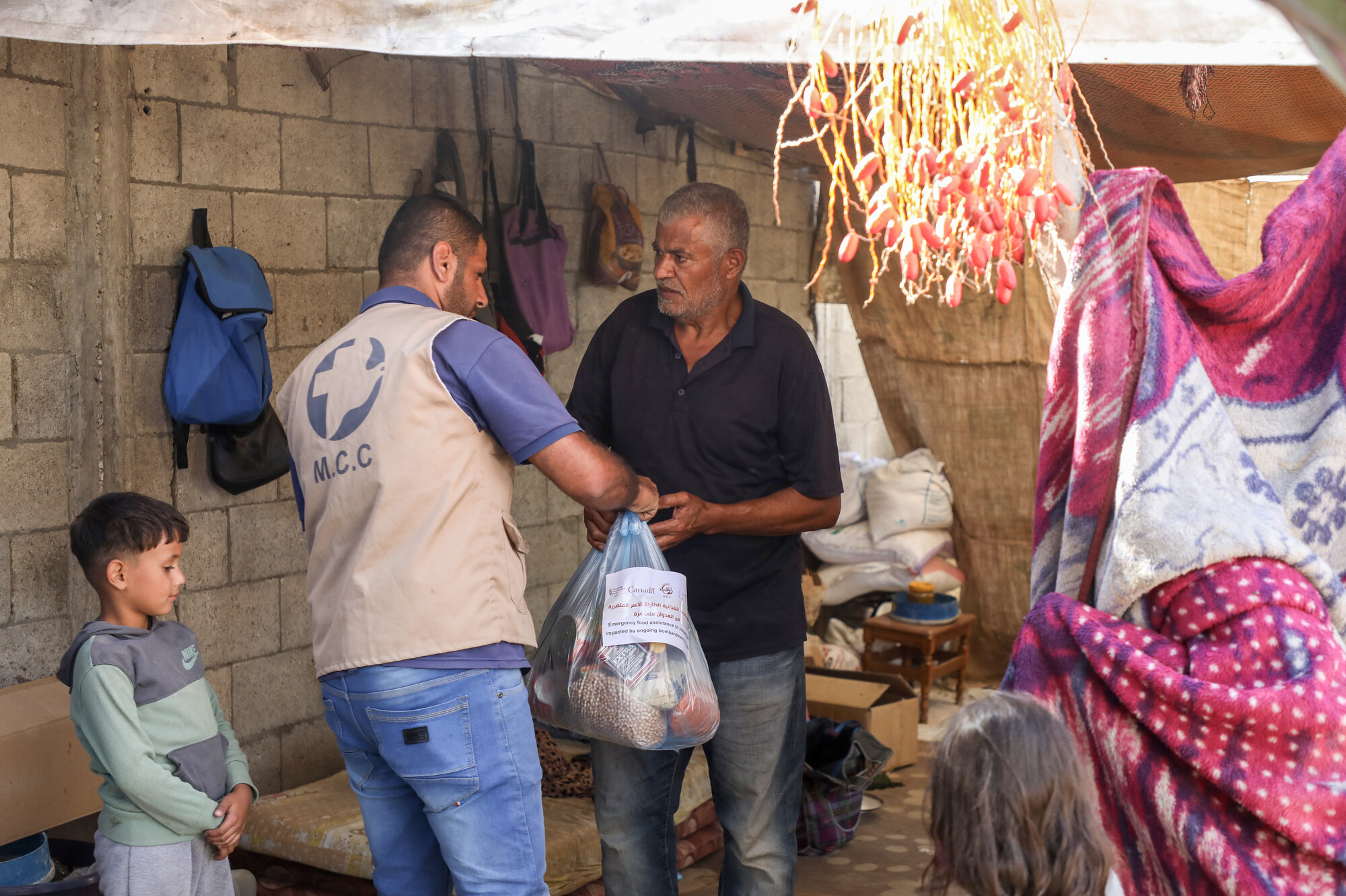 A man wearing an MCC vest gives a man wearing a black shirt a bag of emergency food supply as a child watches the exchange.