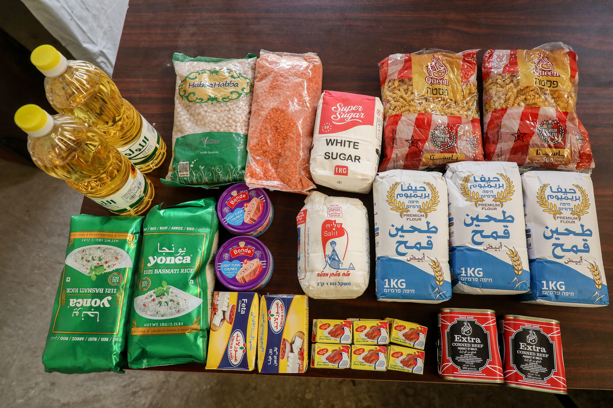 The contents of a food parcel that includes flour, rice, lentils, chickpeas, cooking oil, sugar, canned meat, cheese, tuna and pasta are arrayed on a table.