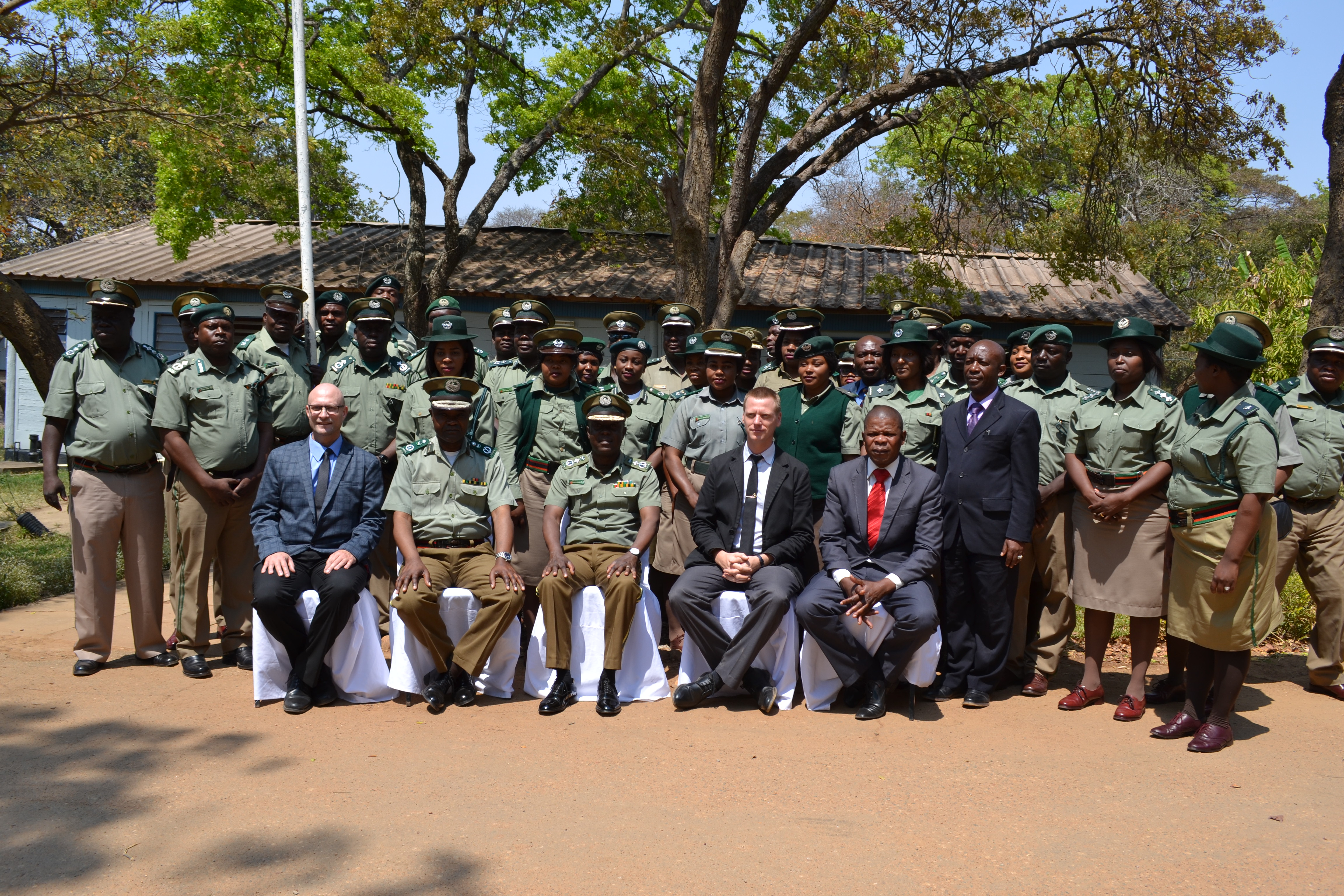 a large group of people, most in police-style uniforms with a seated row of people in suits, face the camera. There are tropical trees in the background