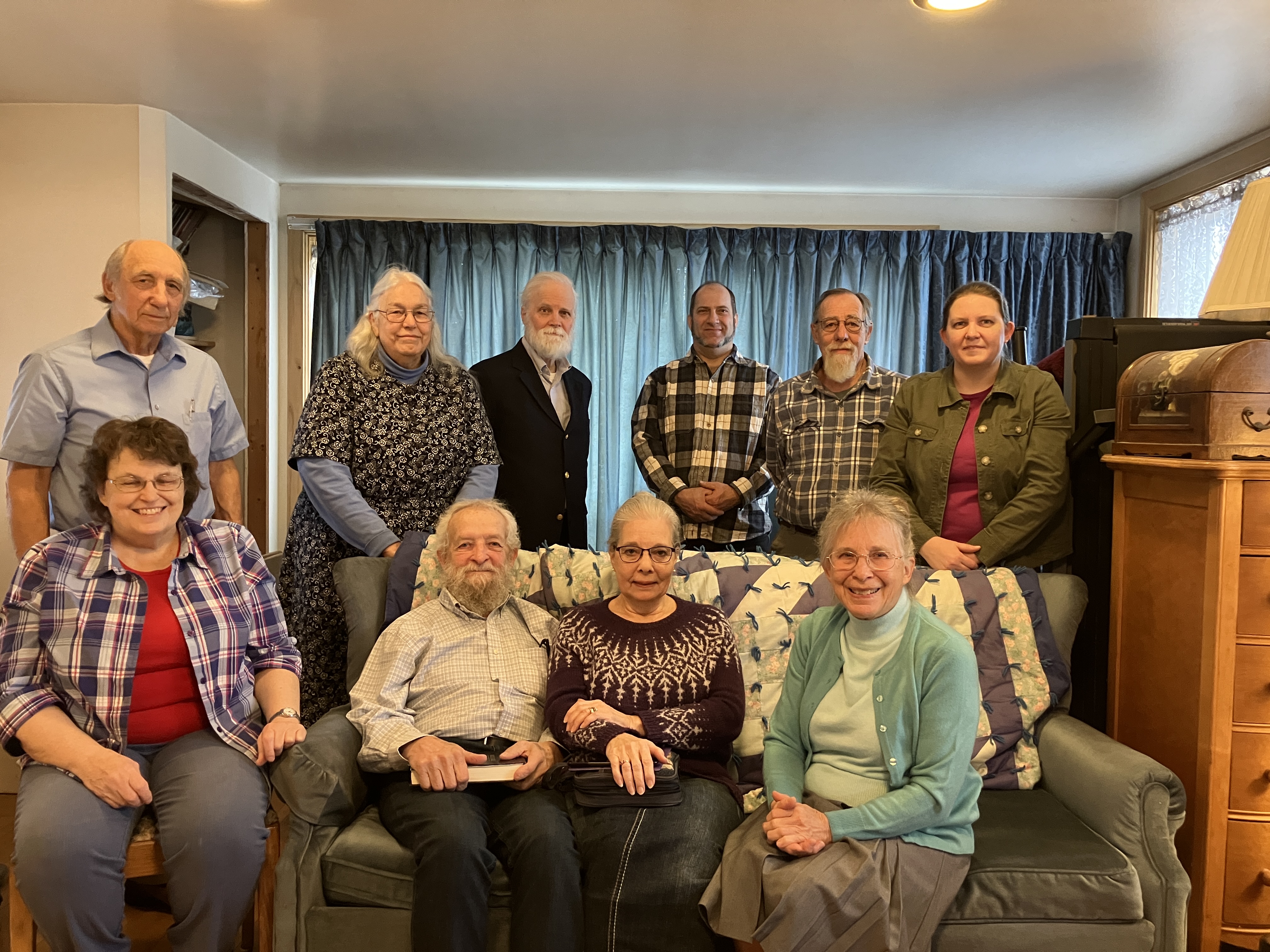 + A group of twelve people poses together in a living room, seated and standing around a couch with various furnishings in the background.
