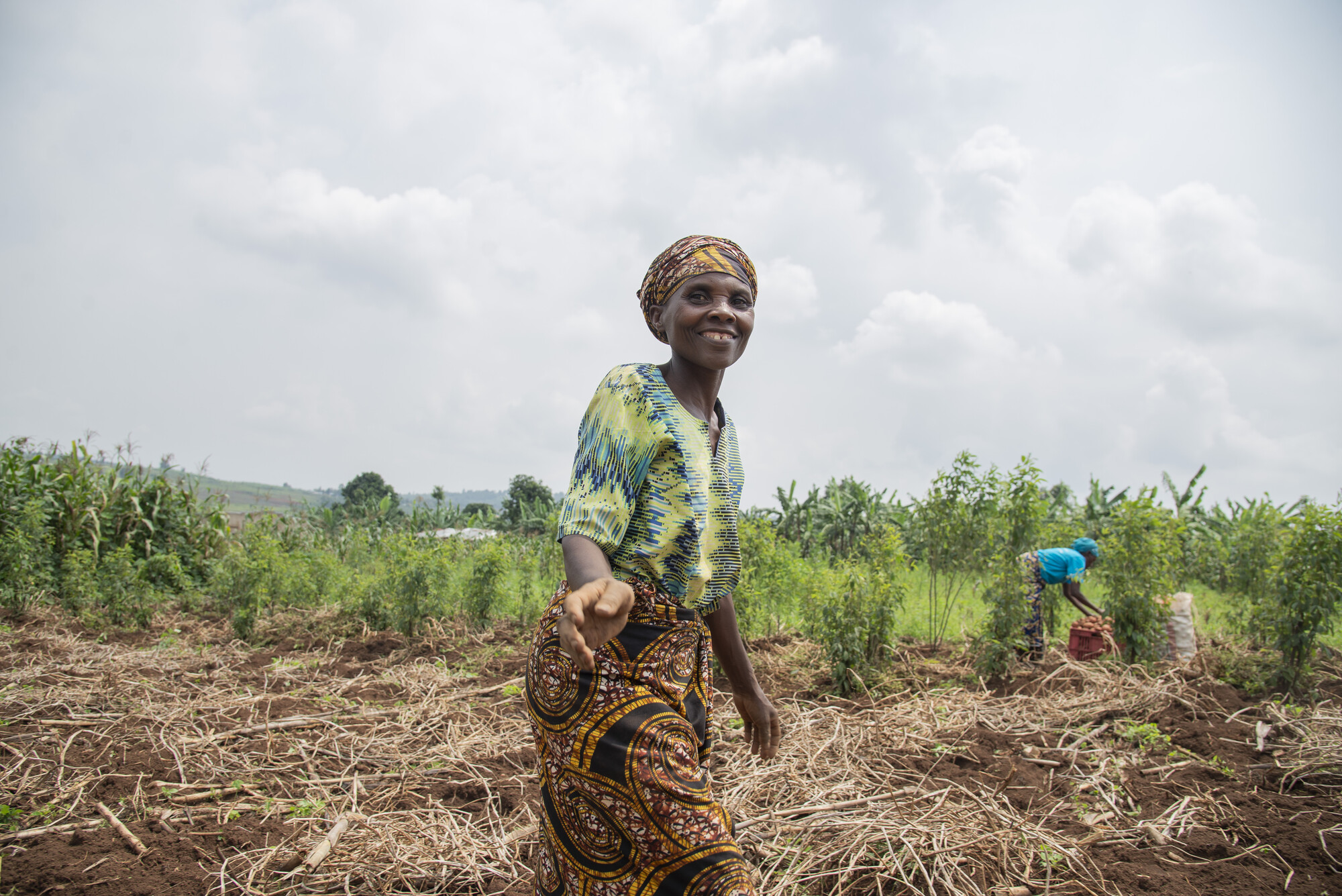 A woman walks in a field under a cloudy sky and looks directly at the camera