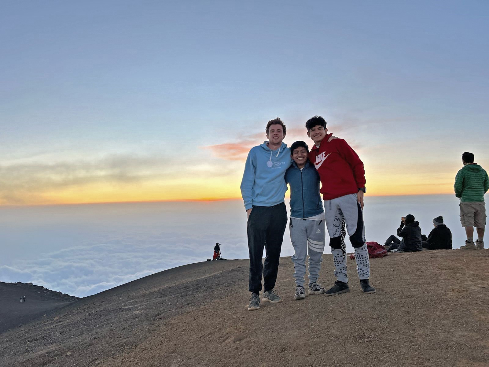 Three people pose together on a mountain at sunset, with a clear sky and clouds below them, suggesting a high altitude. Other individuals are seated in the background.