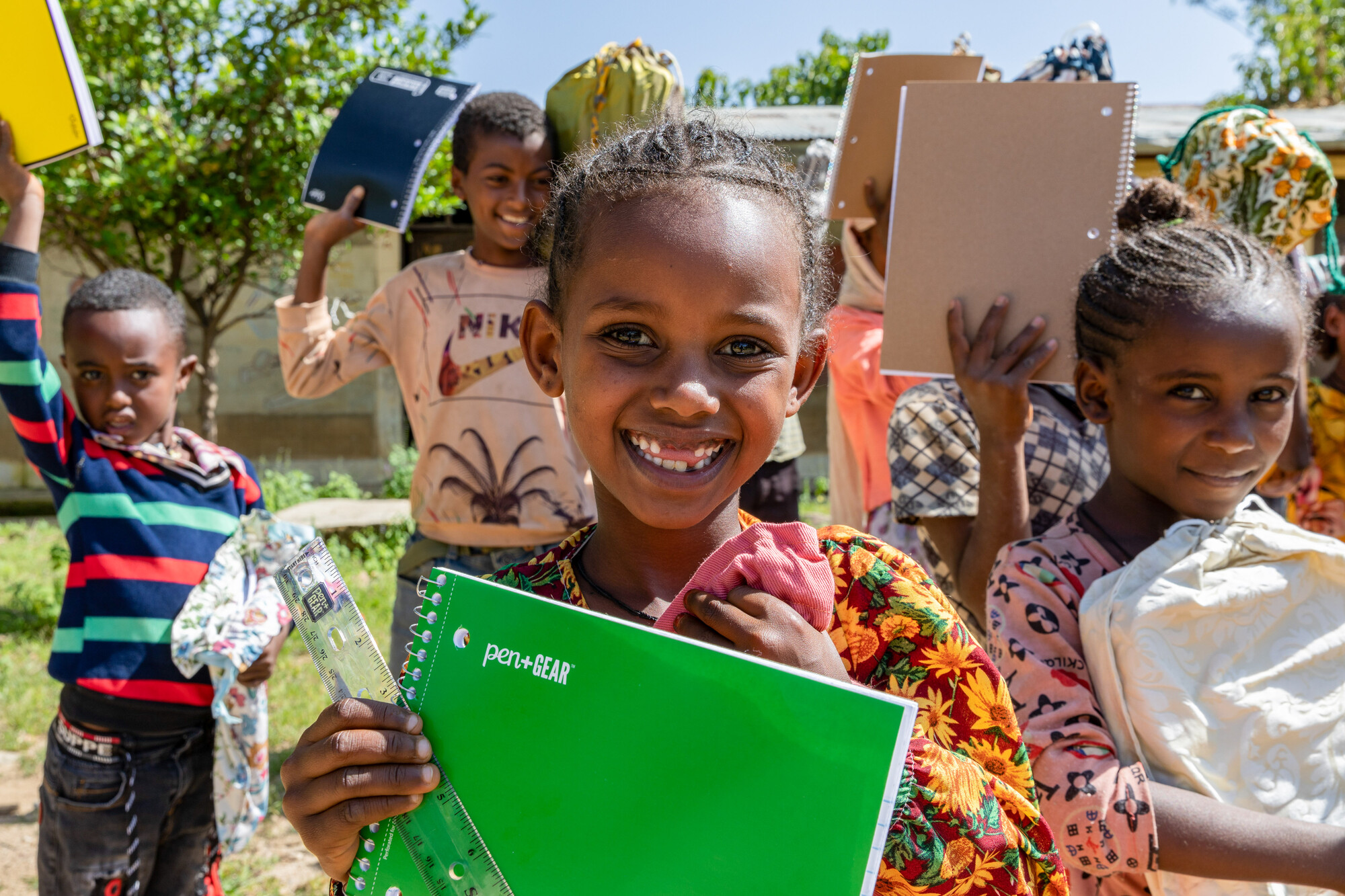 A group of children smiles while holding notebooks and other school supplies, with some raising their items in celebration.