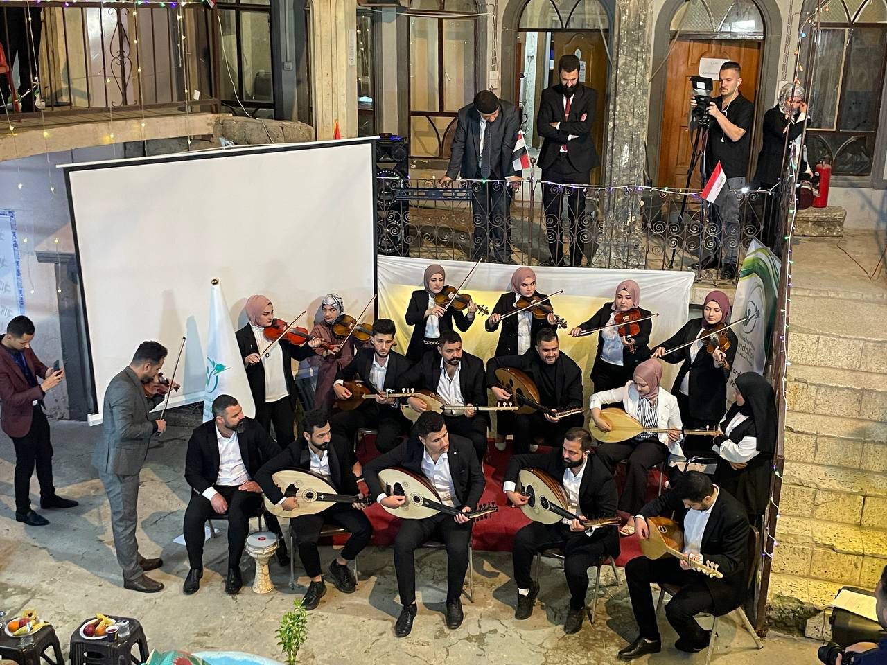 A group of musicians plays instruments on a stage, with some seated and others standing. A screen and audience are visible in the background.