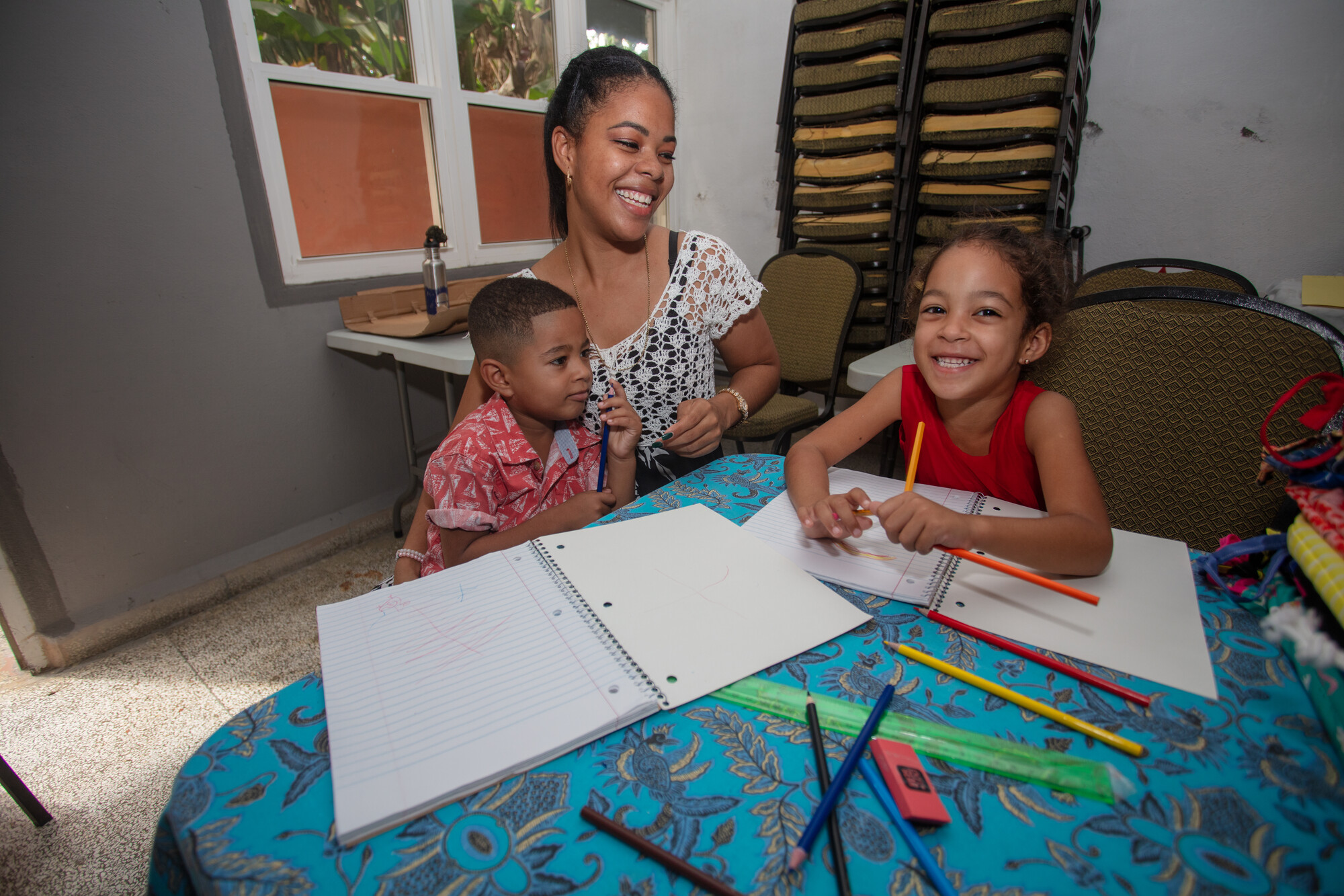 A woman and two children are seated at a table with open notebooks and colored pencils, engaged in drawing and smiling.