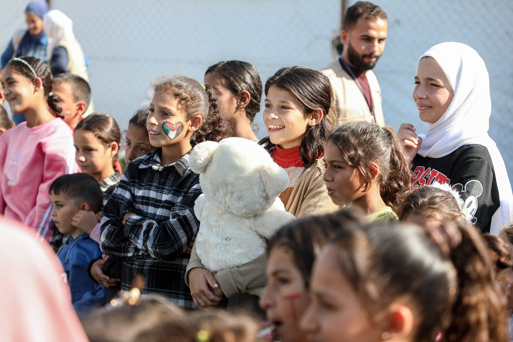 A group of children and adults gather, some smiling, with one girl holding a teddy bear. 