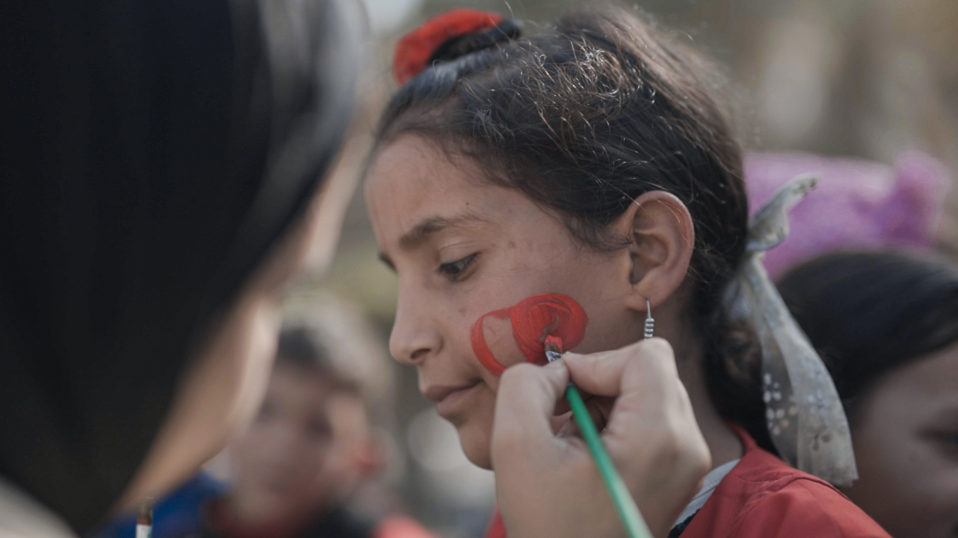 A girl has face paint applied to her cheek while another person holds a brush.