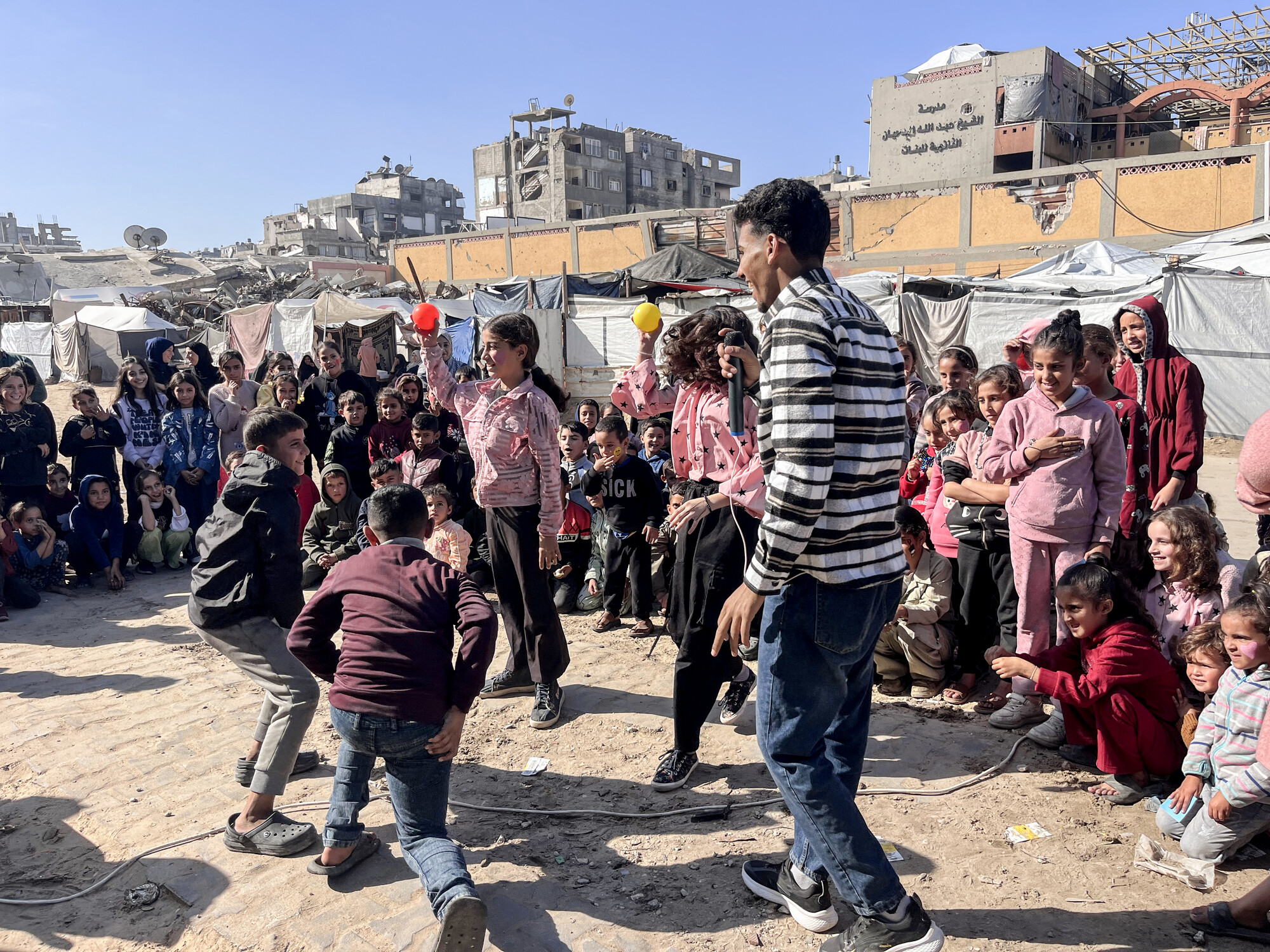 A group of children and adults gather in a lively outdoor setting, watching performers engage in playful activities amidst tents.