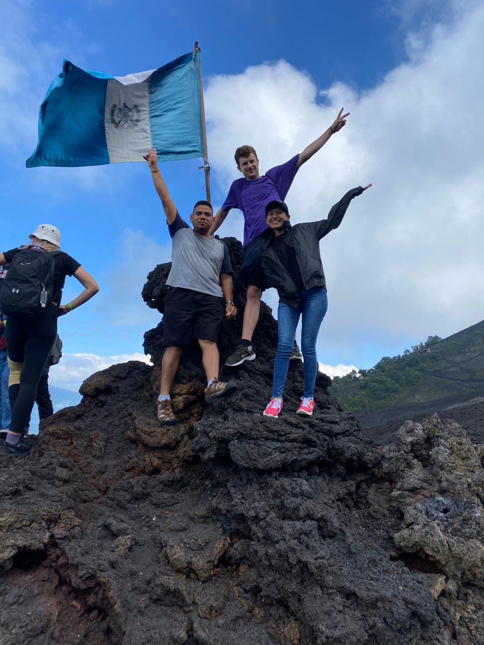 Three individuals stand on a rocky surface, one holding a flag, while others are in the background. The scene is outdoors with a mountainous backdrop.
