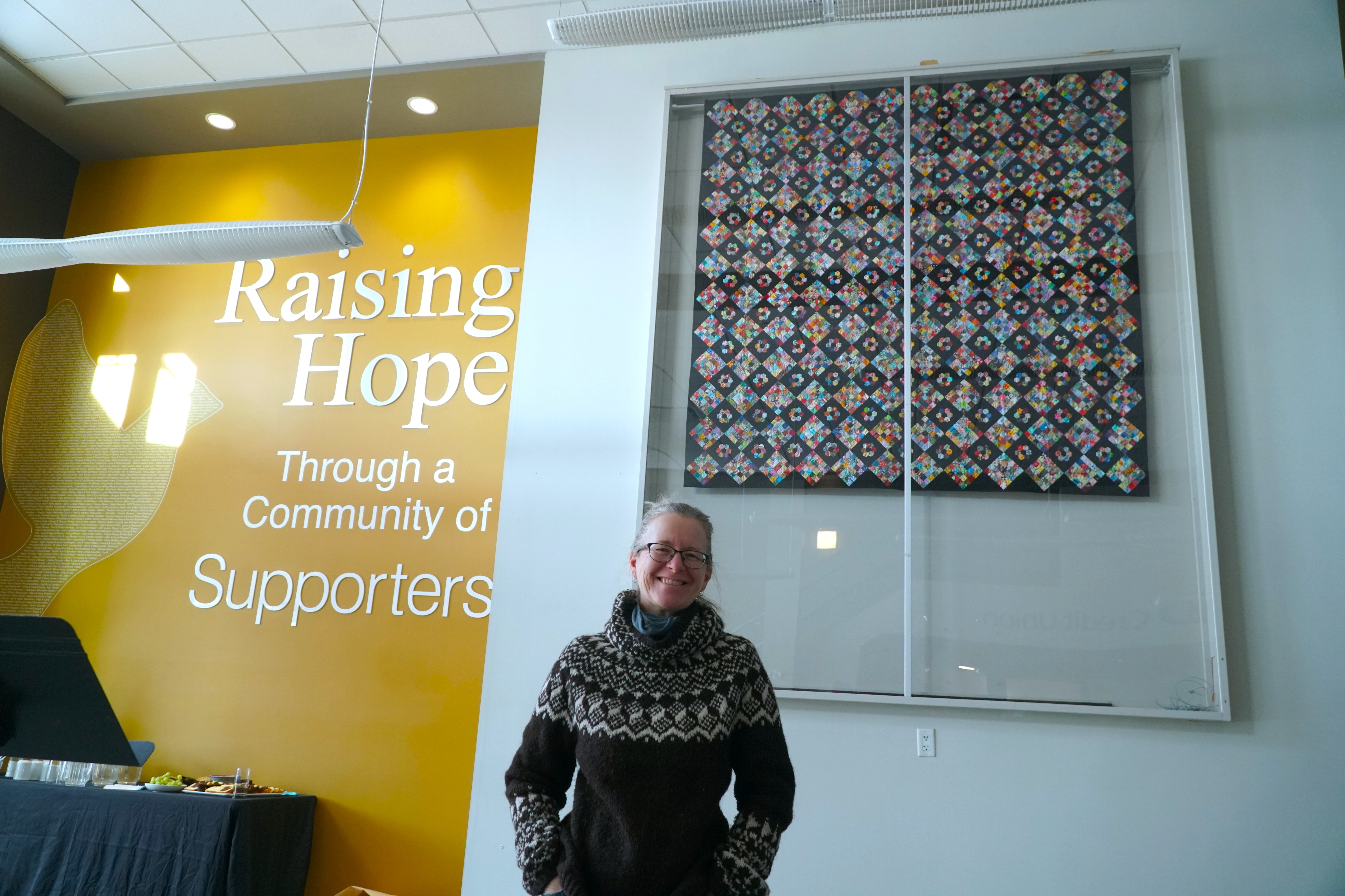 a woman smiles while standing in front of a big quilt hung on the wall