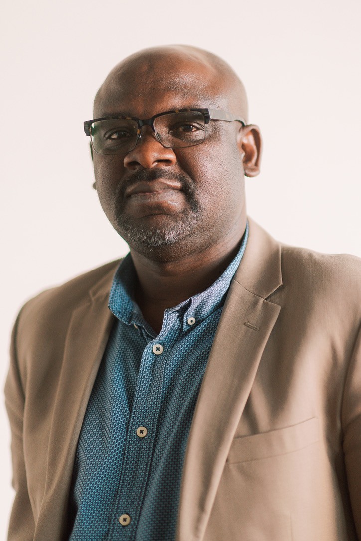 A man with glasses wears a light coloured blazer in a headshot.
