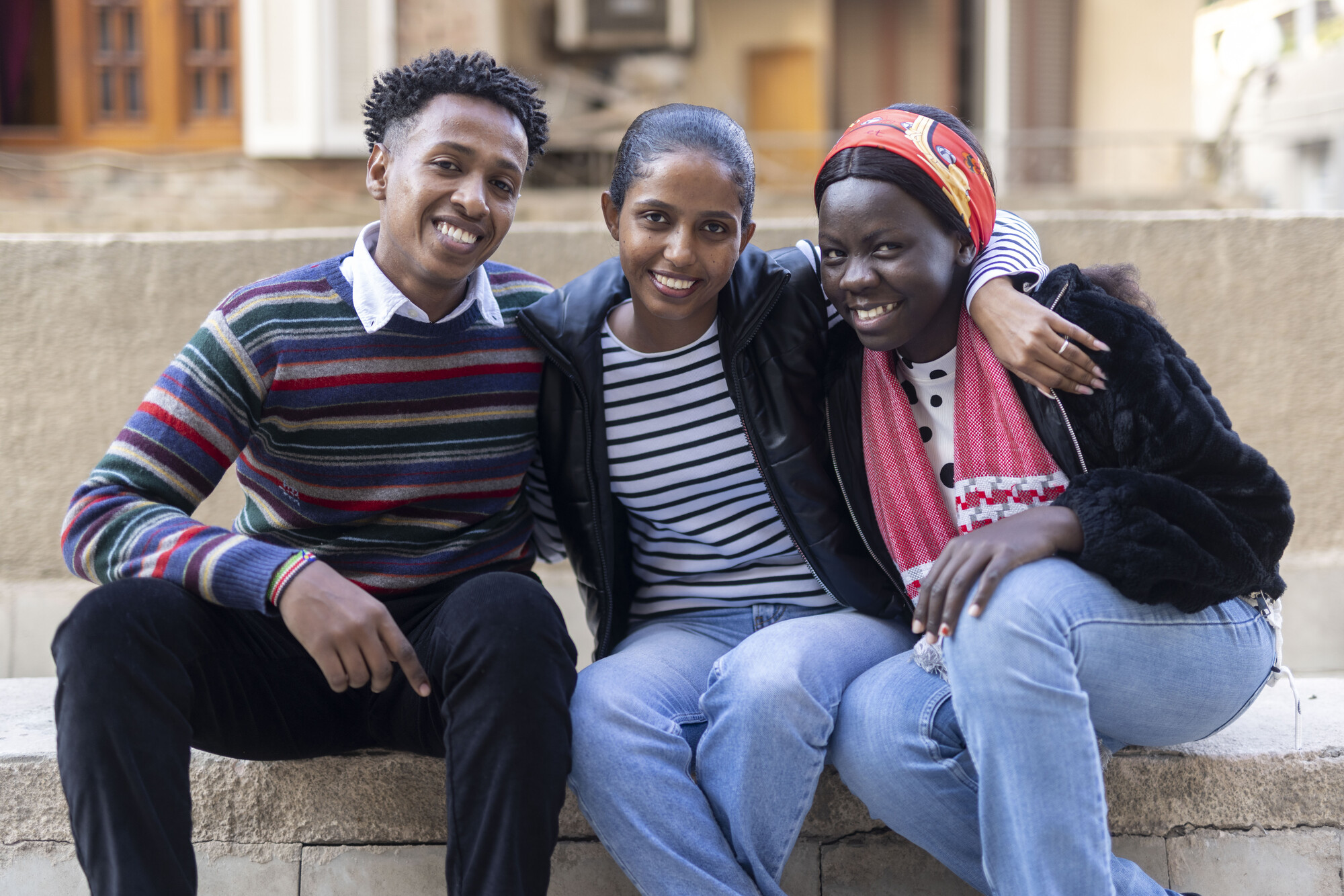 Three individuals sit closely on a stone bench, smiling and posing together. They wear casual clothing and appear relaxed.