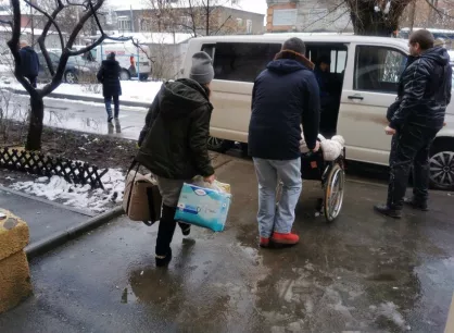 RS115795_KECB-09-scr (1).jpg Three people on a sidewalk are heading towards a white van. One is carrying bags, while one is pushing the other in a wheel chair.