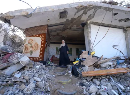 A woman stands amidst rubble and debris, with a partially collapsed building and scattered belongings around her.