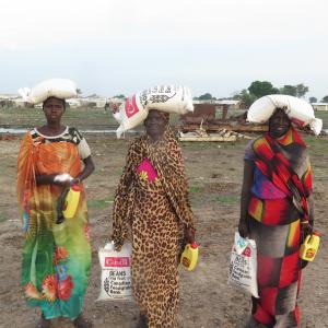 Three South Sudanese woman hold food supplies in their arms and on their heads.