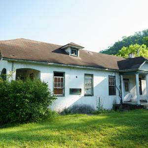 An old, neglected house with peeling white paint, surrounded by greenery, under a clear sky. It appears abandoned and in disrepair, with overgrown bushes.