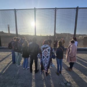 group of people circled in front of border wall between the US and Mexico