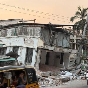 A family on a moped pass a building that's collapsed due to earthquake damage.