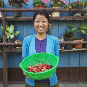 A smiling person presents a colander of strawberries. There are rows of potted plants in the background.