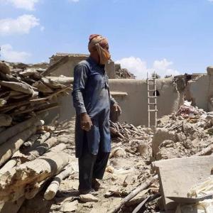 A person stands amidst the rubble and debris that was his house, with a ladder leaning against a damaged wall and remnants of structures around.