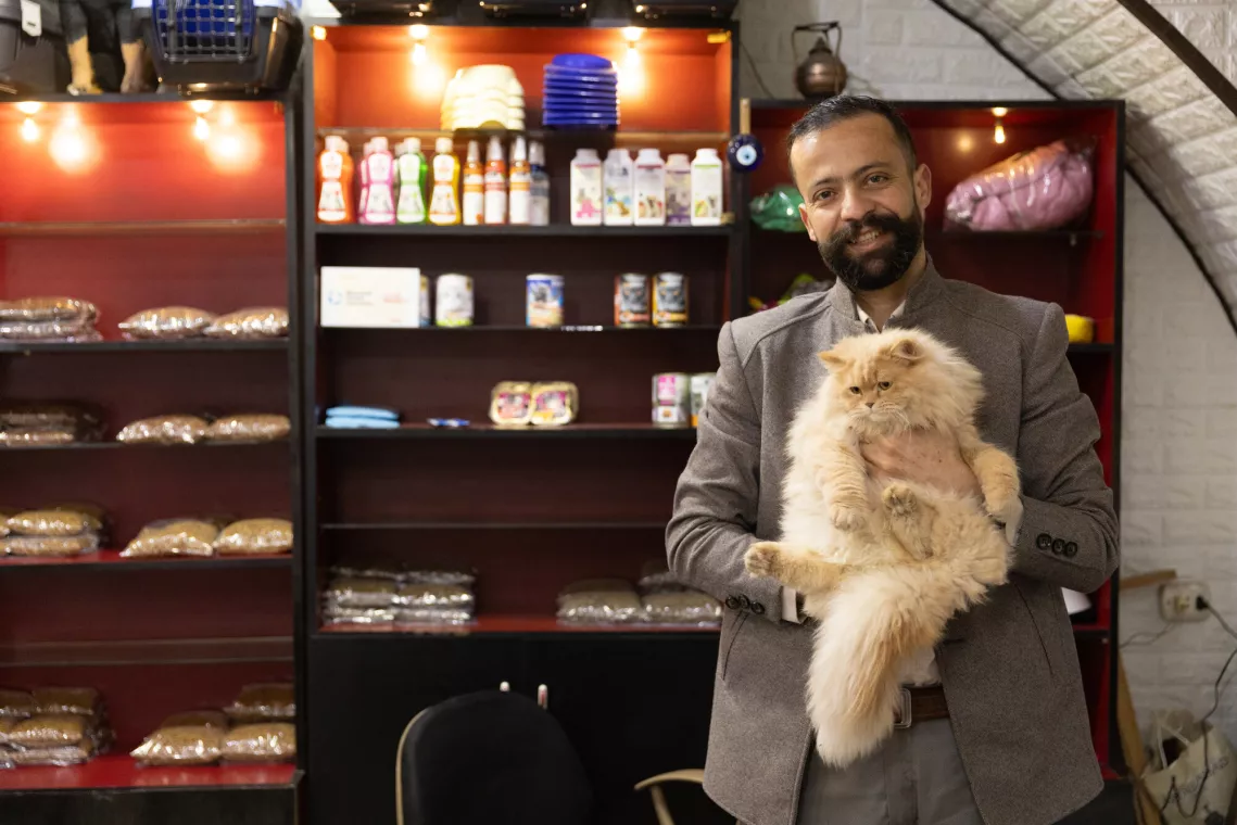 A man in a blazer smiles while holding a fluffy cat, standing in a room with shelves stocked with various items, including bread and beverages.